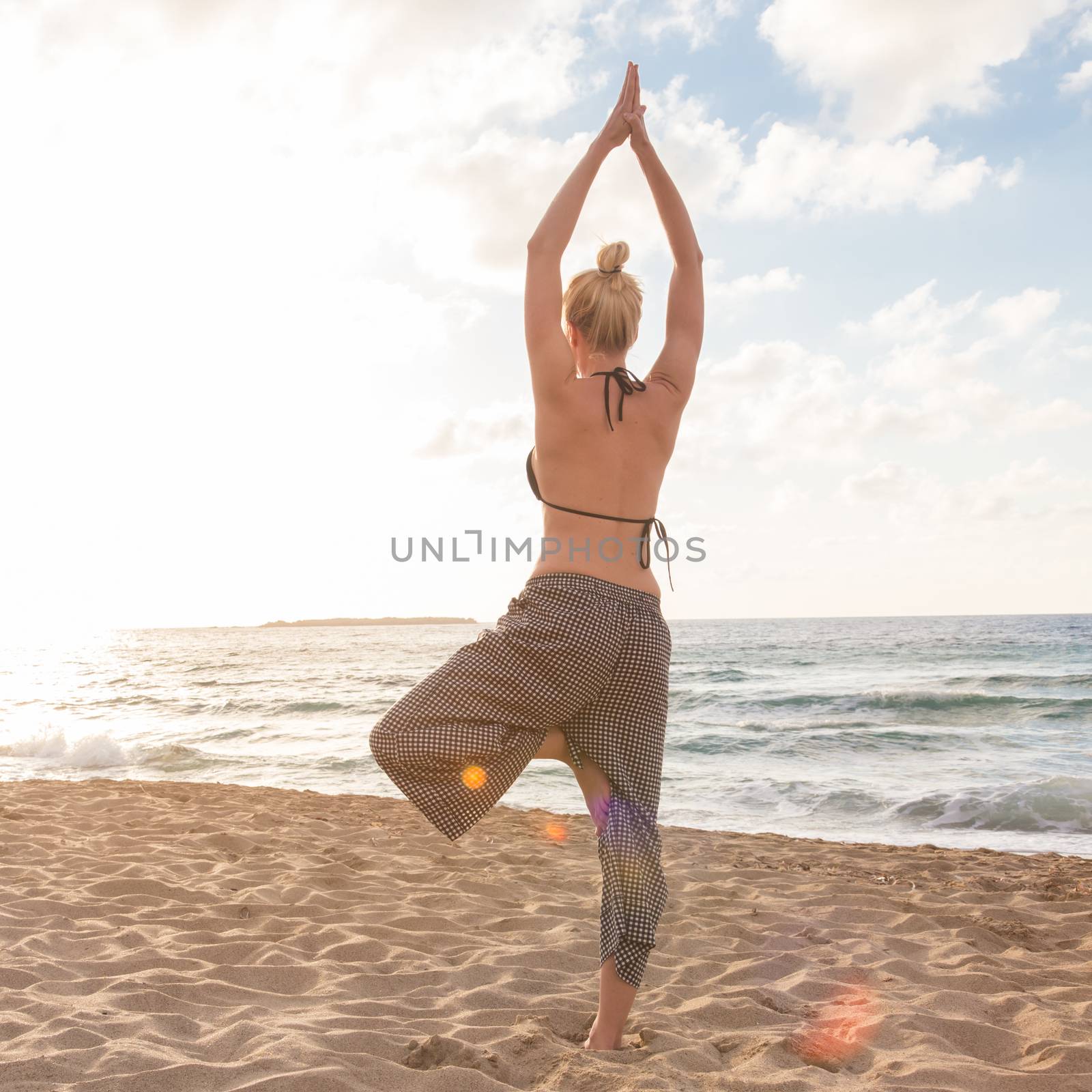 Silhouette of young woman practicing standing tree yoga pose on sandy beach at sunset.