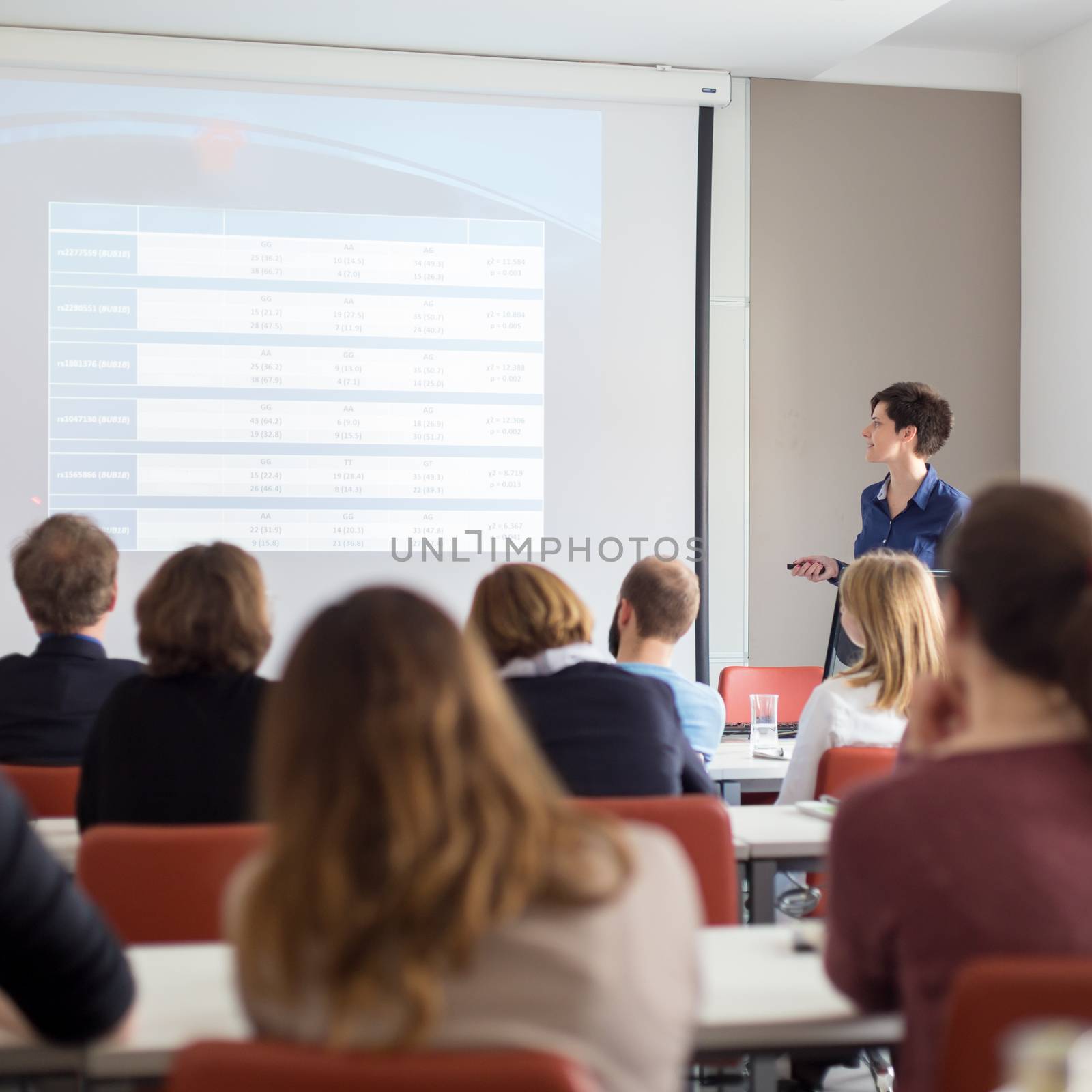 Female speaker giving presentation in lecture hall at university workshop . Participants listening to lecture and making notes. Scientific conference event.