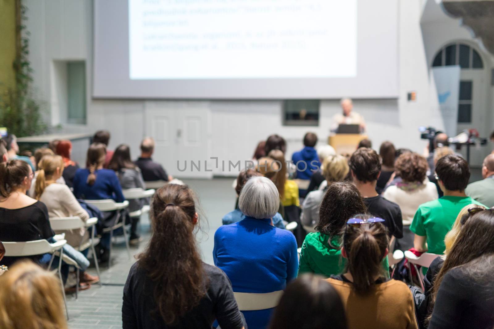 Male speaker giving presentation in lecture hall at university workshop. Audience in conference hall. Rear view of unrecognized participant in audience. Scientific conference event.