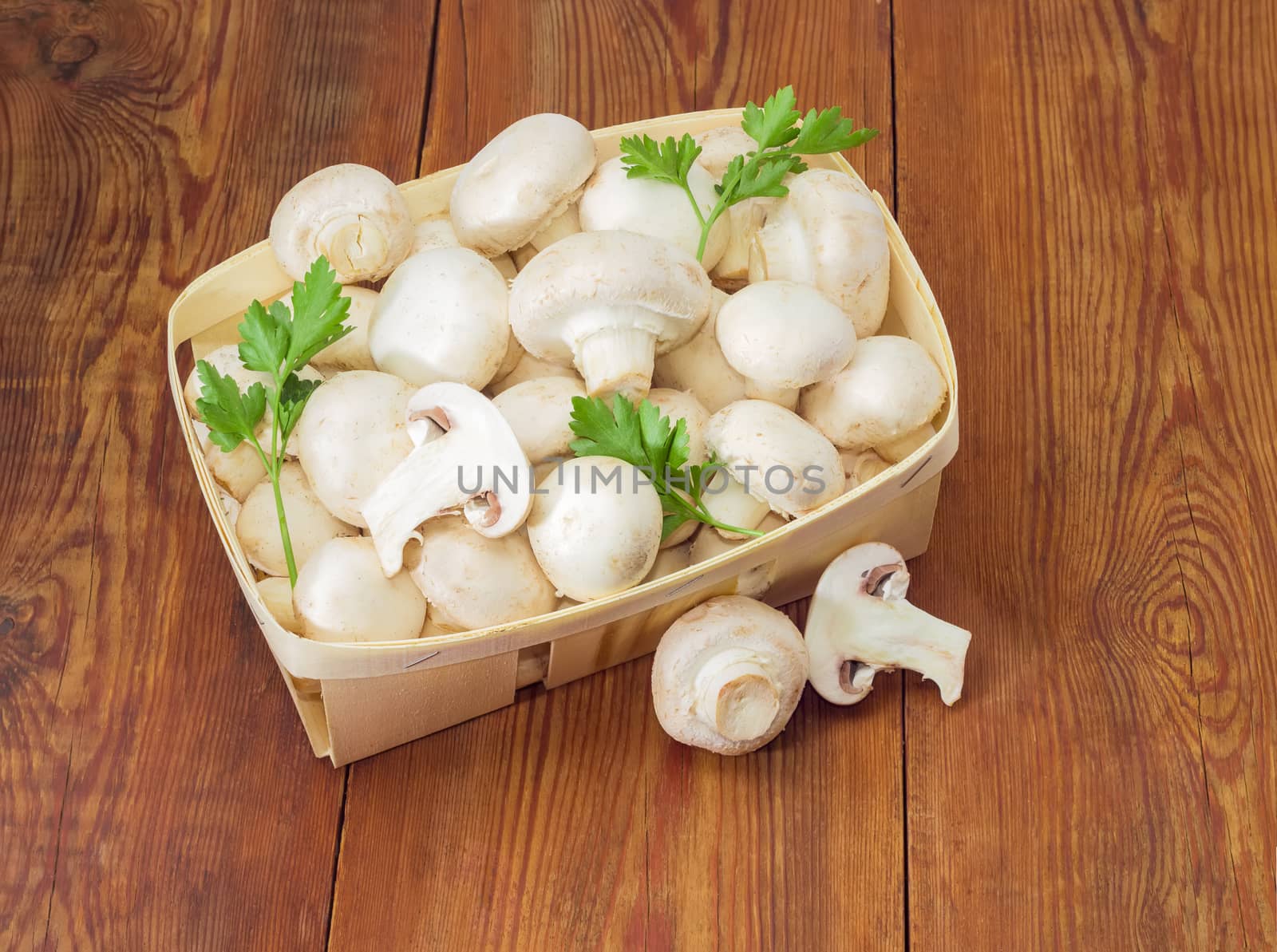 Fresh cultivated button mushrooms and twigs of parsley in the wooden basket, one whole mushroom and mushroom cut in half separately beside on an old wooden surface
