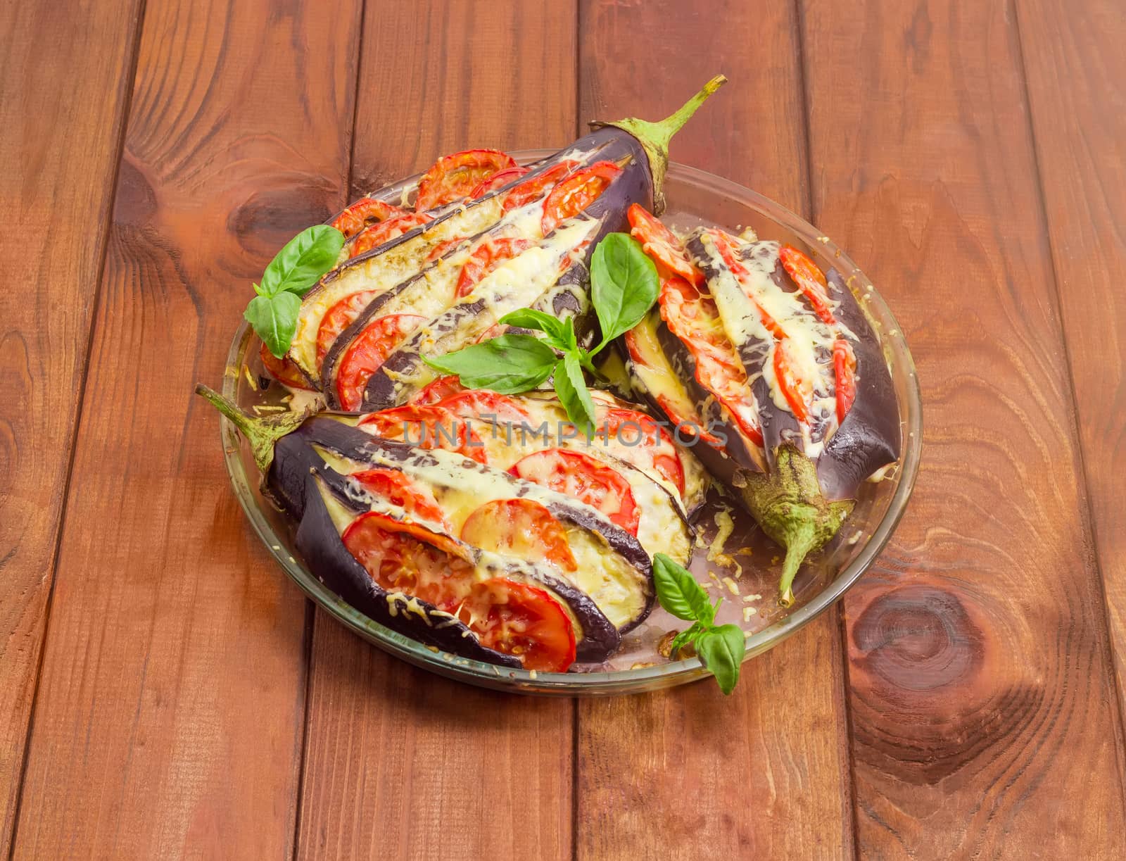 Three baked eggplants stuffed with vegetables and cheese in round glass baking tray on a surface of dark wooden planks
