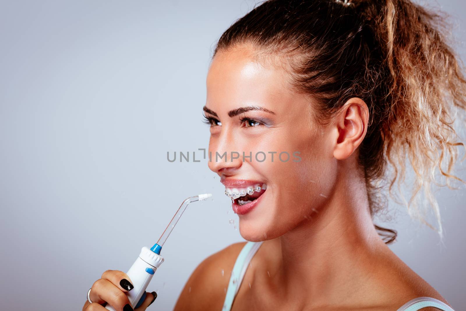Smiling young woman with braces cleaning her teeth with oral irrigator.