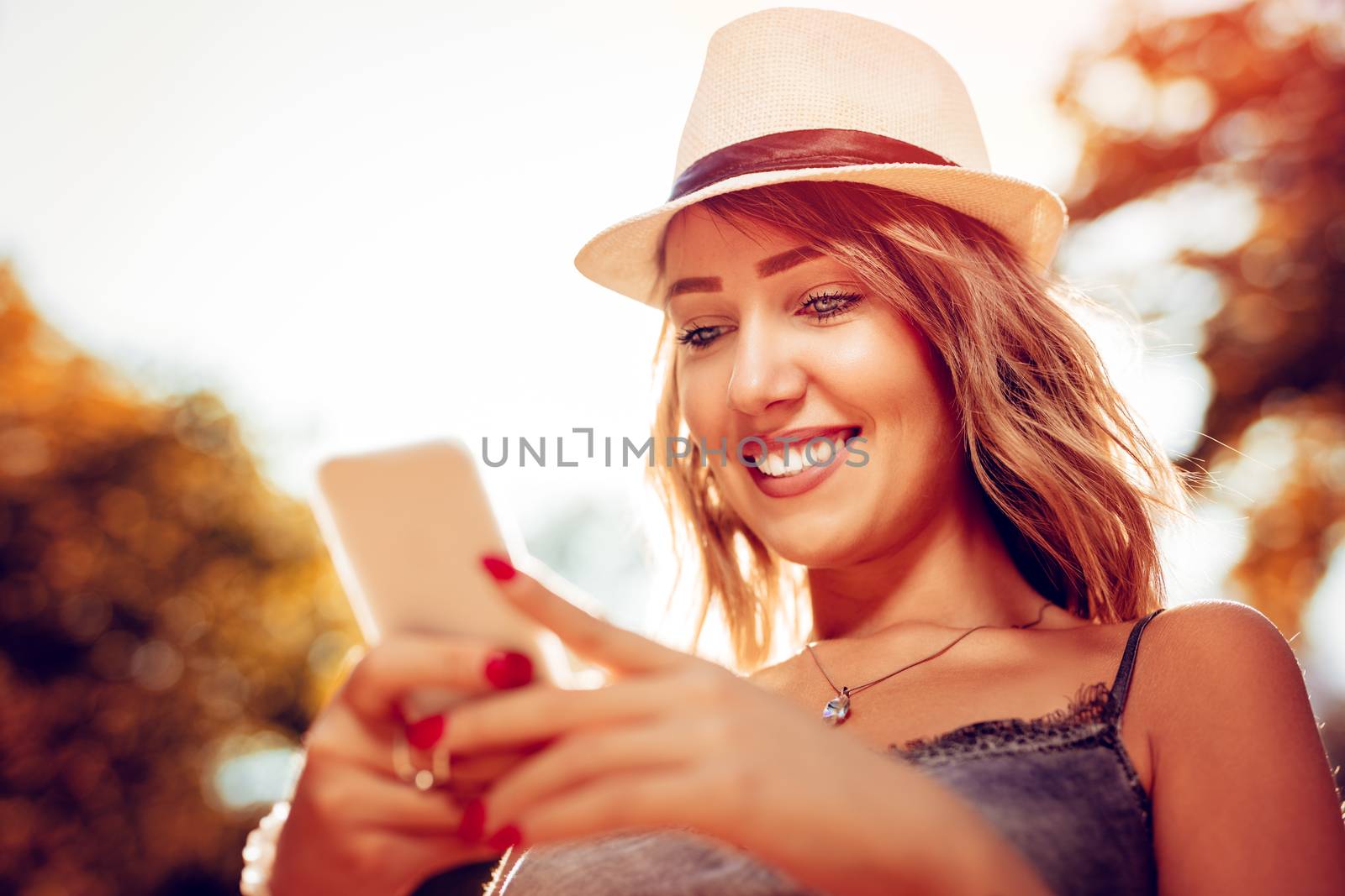 Beautiful smiling young woman with summer hat enjoying in the nature in autumn and using smartphone.