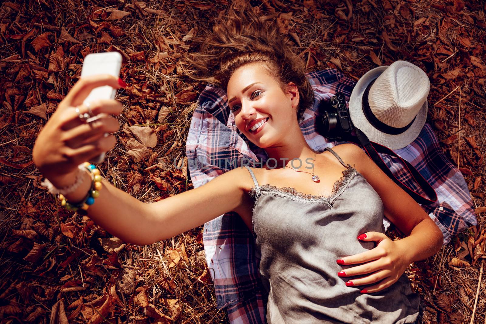 Beautiful smiling girl taking selfie in nature in autumn. She is lying on the falls leaves.
