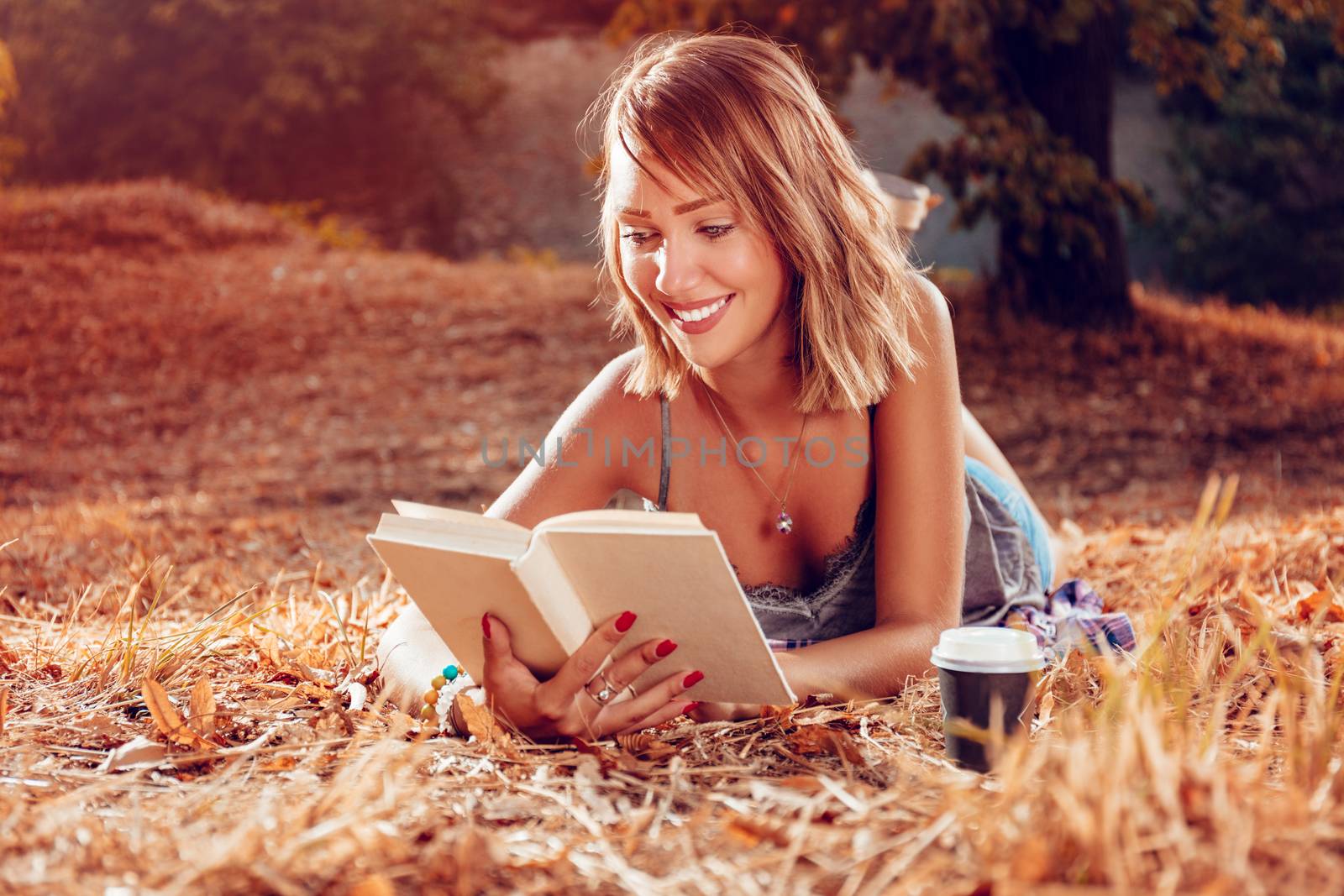 Cute smiling young woman lying on the withered grasses in autumn and reading book.