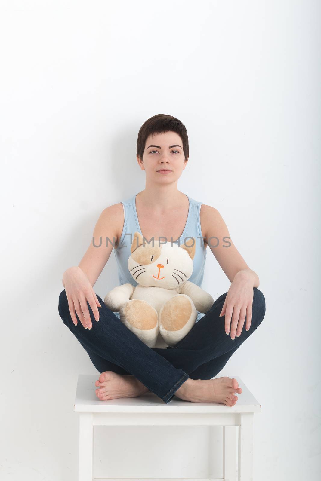 Young attractive smiling woman practicing yoga, sitting in Half Lotus exercise Ardha Padmasana pose, working out wearing dark bleu jeans indoor full length, white plush toy cat on her lap.