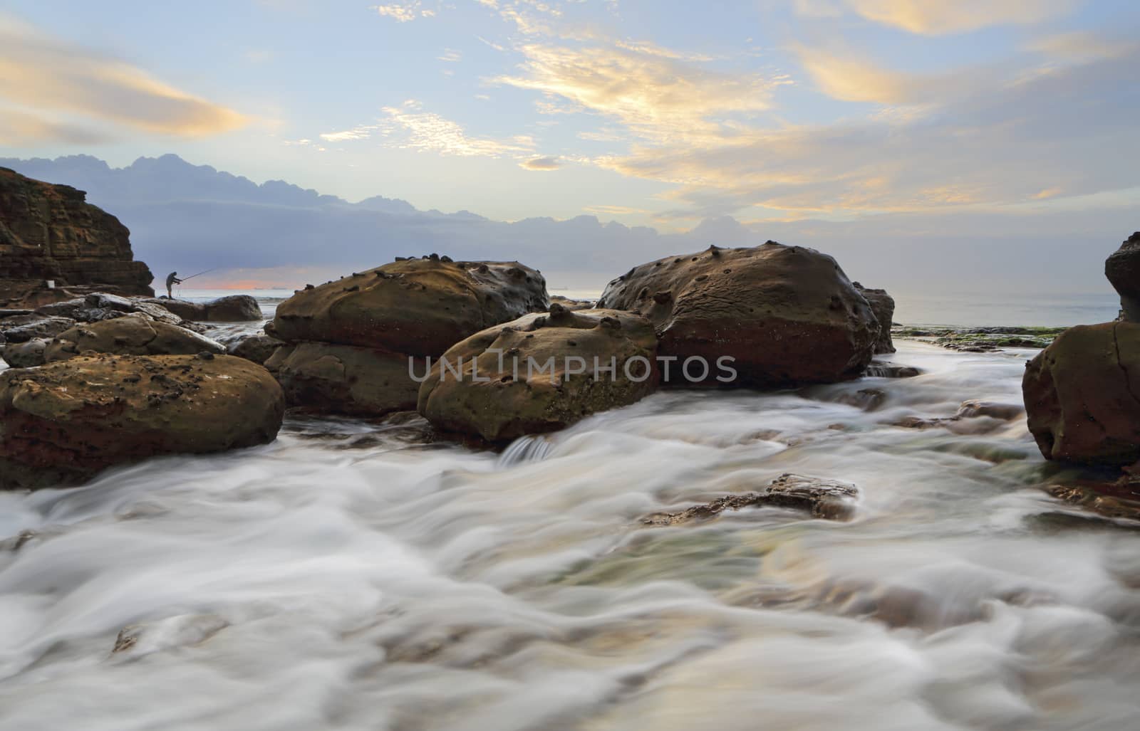 Strong rock flows as large waves wash up onto the rock shelf and then cascade rapidly down creating beautiful motion. The rocks here hve strange wart like nodules.   A rock fisherman in the distance.  Location Wollongong