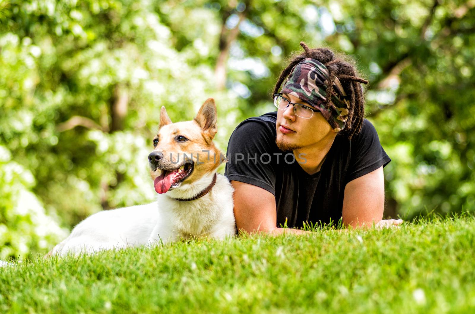 young man with dreadlocks is laying on the grass with his dog by Desperada