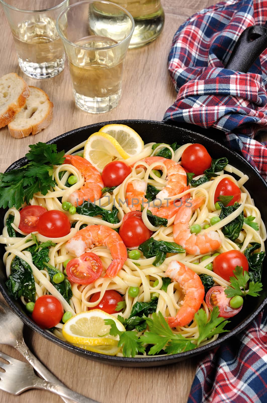 Pasta with fried prawns, peas, tomatoes and spinach in a frying pan, on a table with cider glasses.
