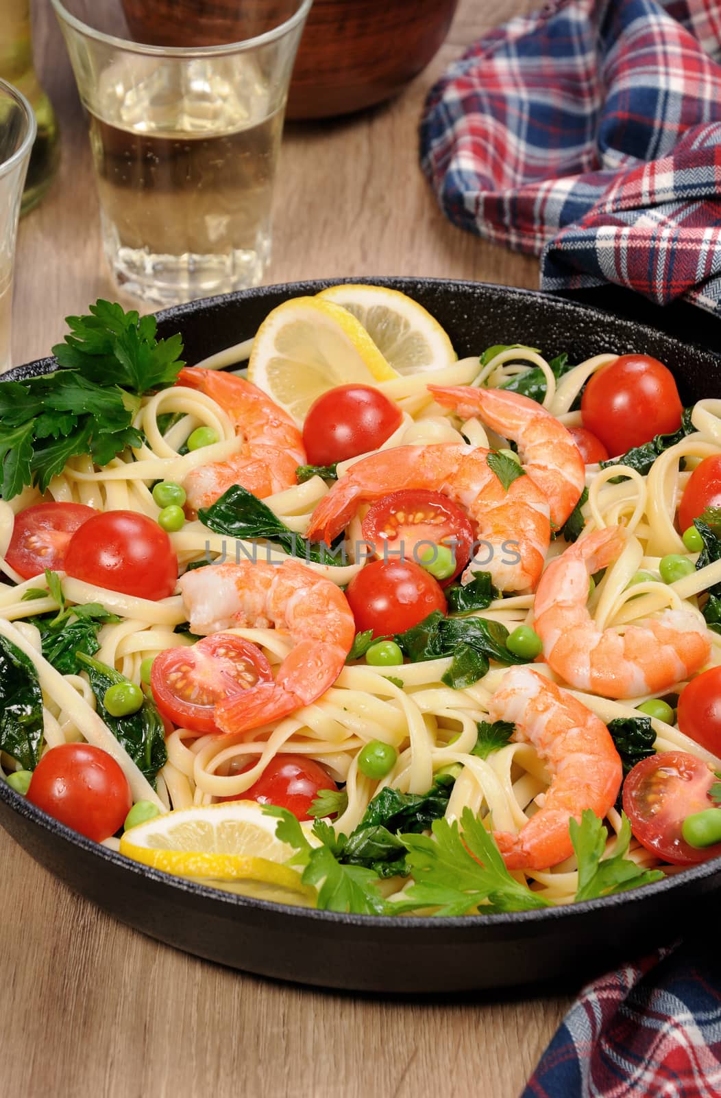 Pasta with fried prawns, peas, tomatoes and spinach in a frying pan, on a table with cider glasses.