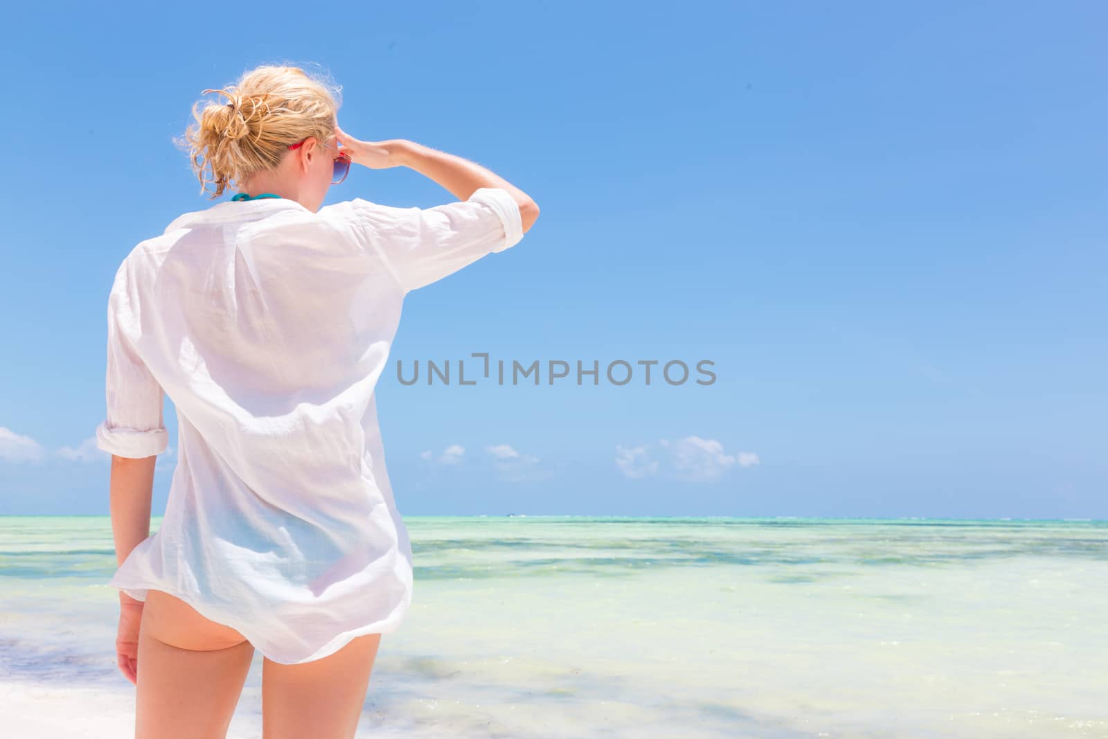 Happy woman enjoying, relaxing joyfully in summer by tropical blue water. Beautiful caucasian model wearing white beach tunic on vacations looking down picture perfect Paje beach, Zanzibar, Tanzania.