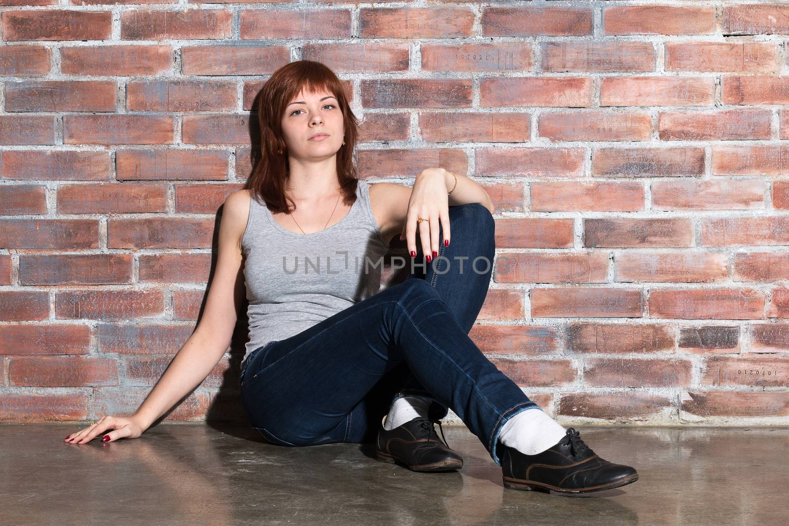 Attractive young woman in casual clothes, blue jeans and vest grey is looking at camera and smiling sitting on a floor by the brick wall. Hipster girl. Youth fashion, beauty. Red hair, haircare. Horizontal.