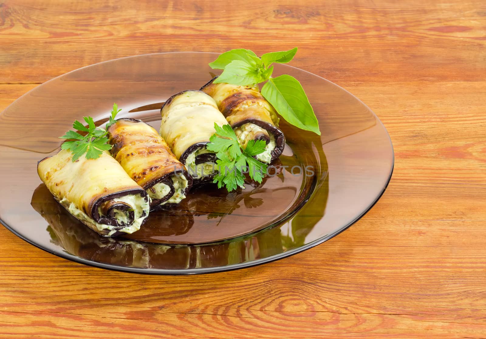 Fragment of a dark glass dish with several eggplant rolls stuffed with tuna and processed cheese and decorated with parsley and basil twigs on a surface of an old wooden planks
