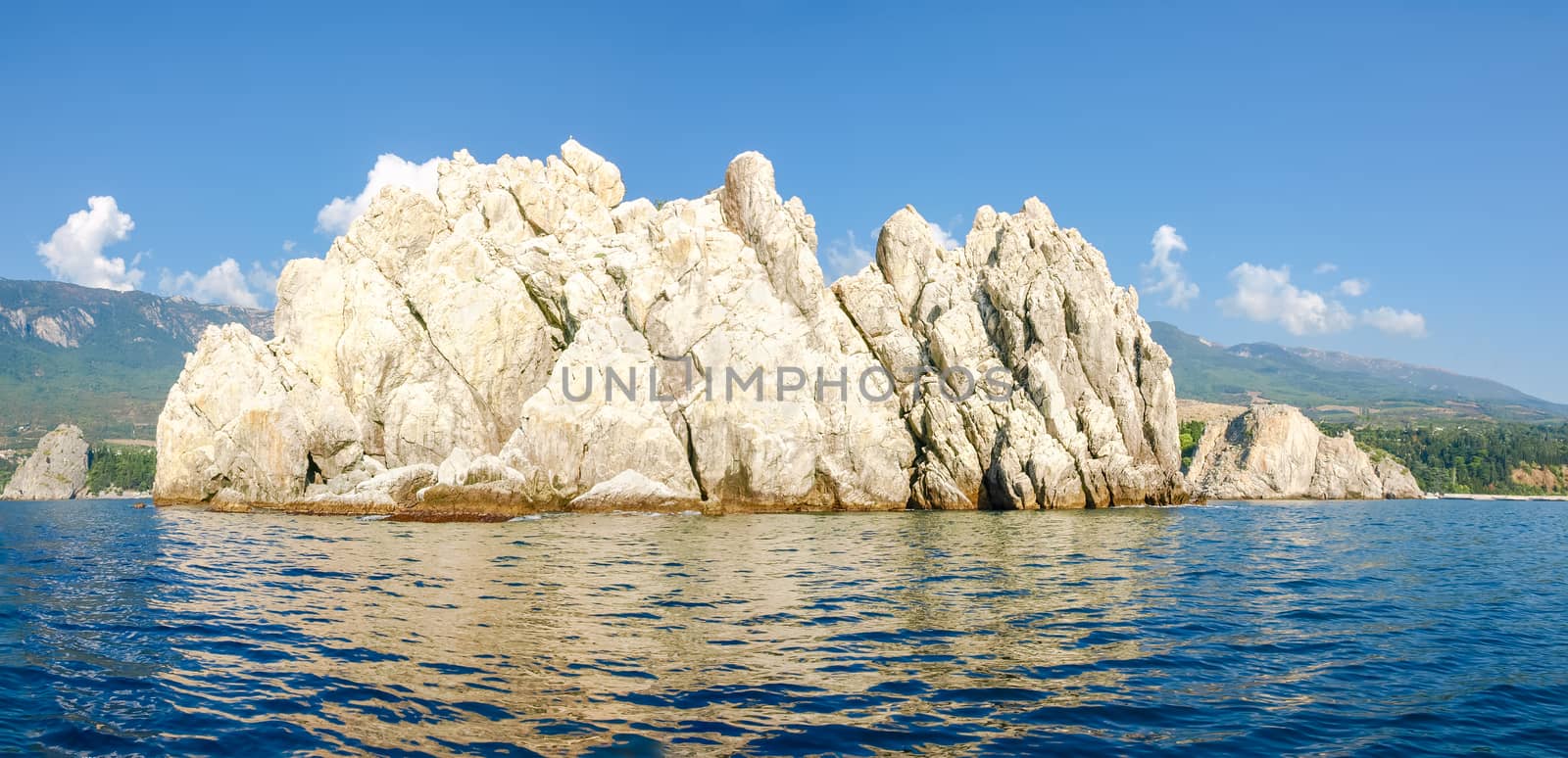 Panorama of the single limestone rock in the sea on a background of the sky and shore with rocks and mountain slopes overgrown with forest
