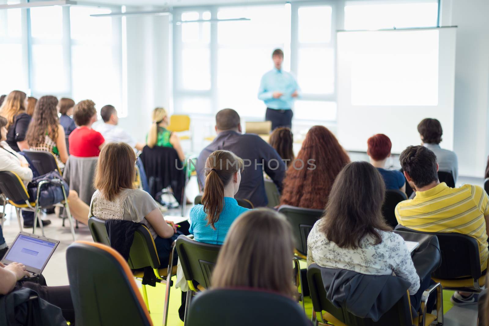 Business and entrepreneurship symposium. Speaker giving a talk at business meeting. Audience in conference hall. Rear view of unrecognized participant in audience.