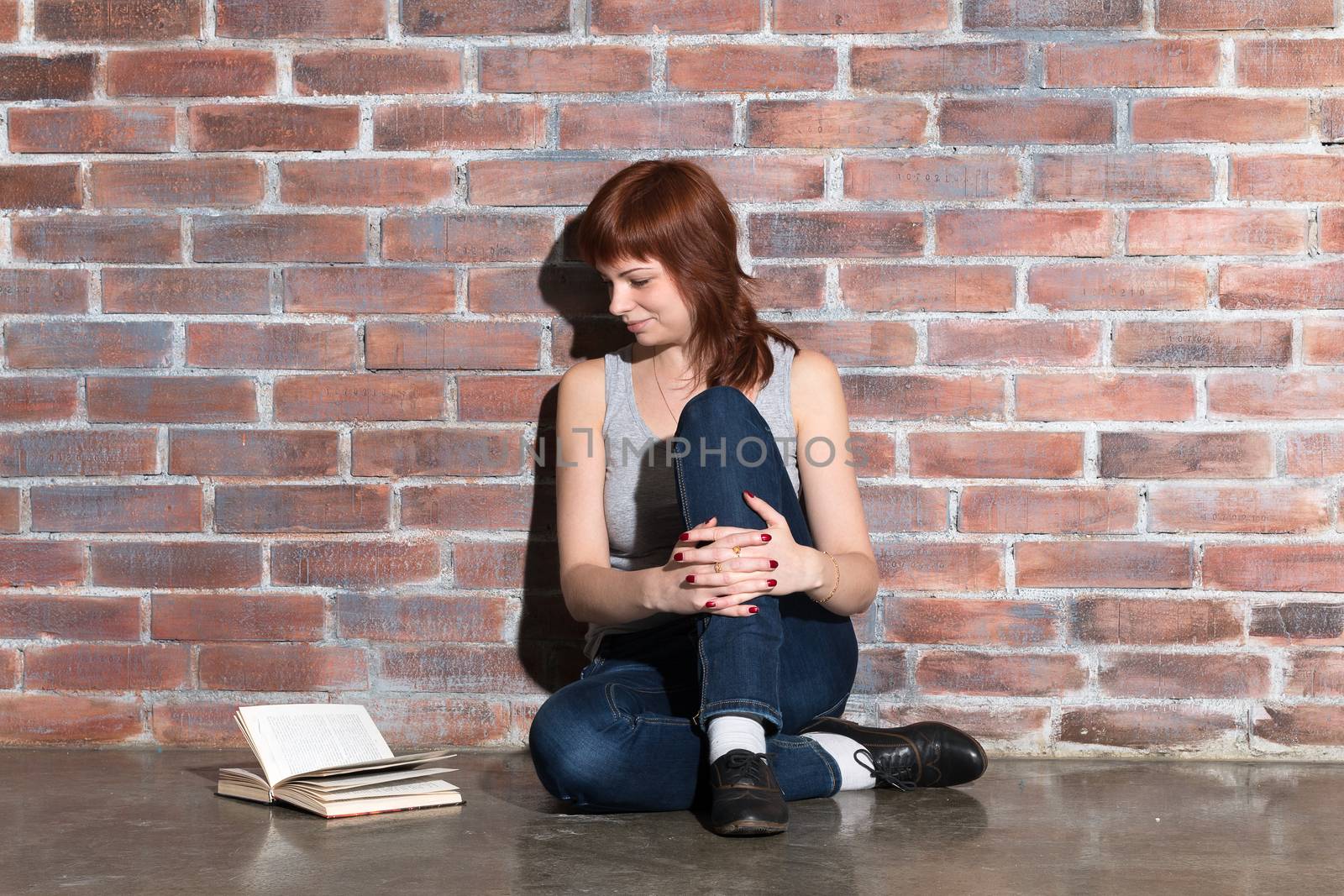 Young beautiful ginger hair woman in blue jeans reading a book while sitting on the floor near red brick wall