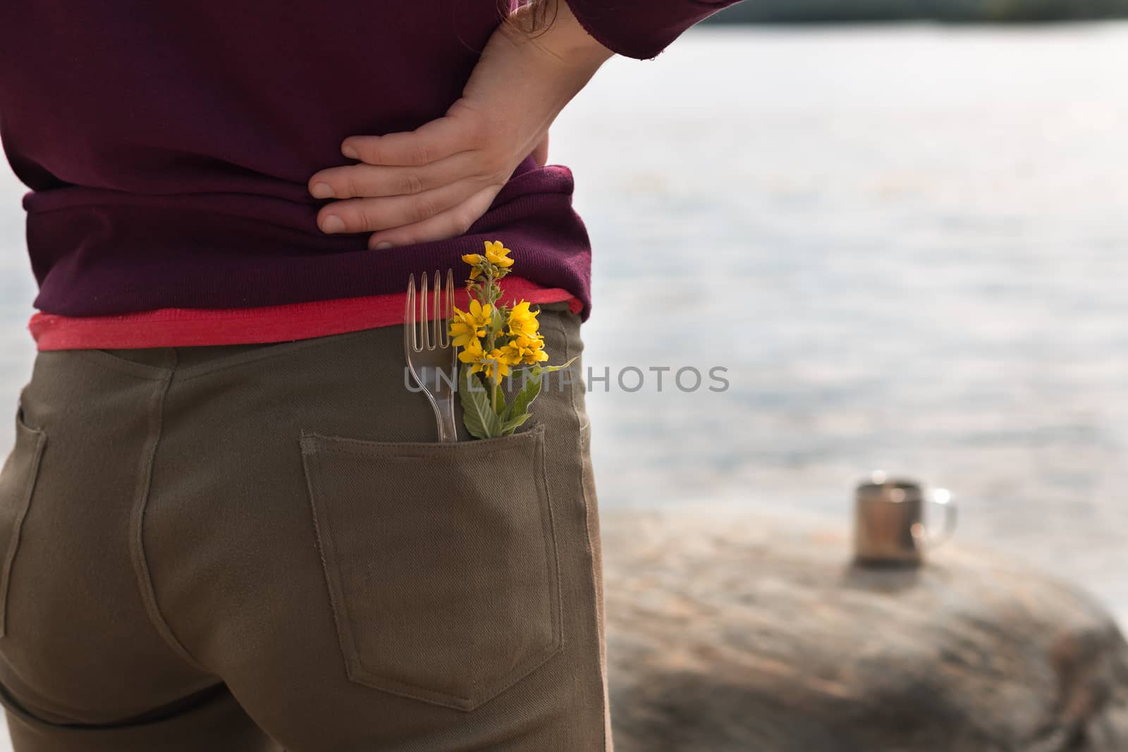 Handsome female tourist wearing brown jeans and purple sweatshirt with fork and yellow flowers in her pocket stands on river bank and enjoys view of mountains in summer. Hike or trip concept. Horizontal