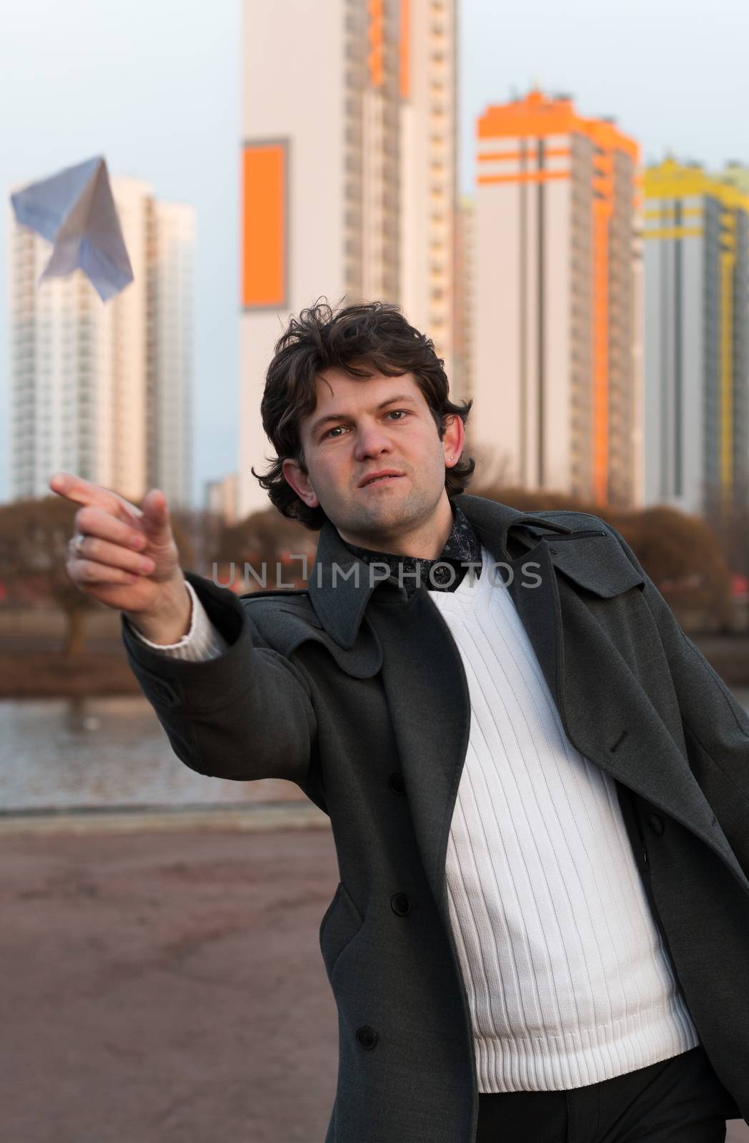 Paper airplane launching in park, free space, Man in grey coat and white sweater holding handmade paper airplane in hand on high-rise building background. Open-air game, rest, leisure, pastime concept.