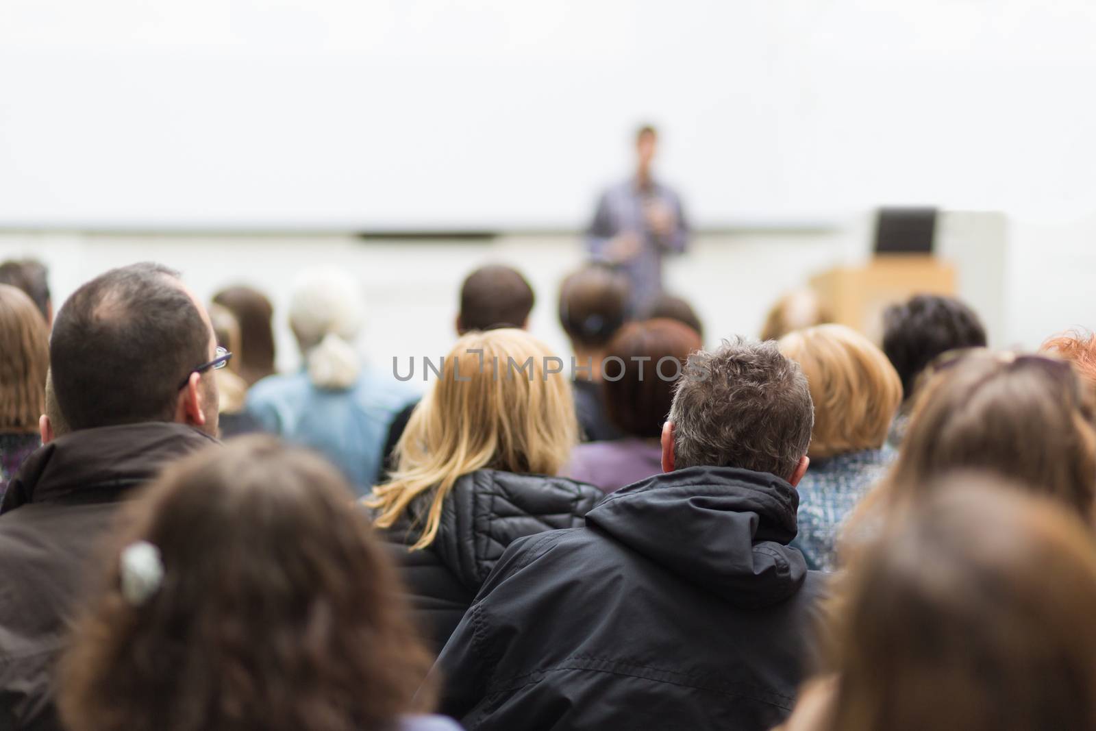 Male speaker giving presentation in lecture hall at university workshop. Audience in conference hall. Rear view of unrecognized participant in audience. Scientific conference event.
