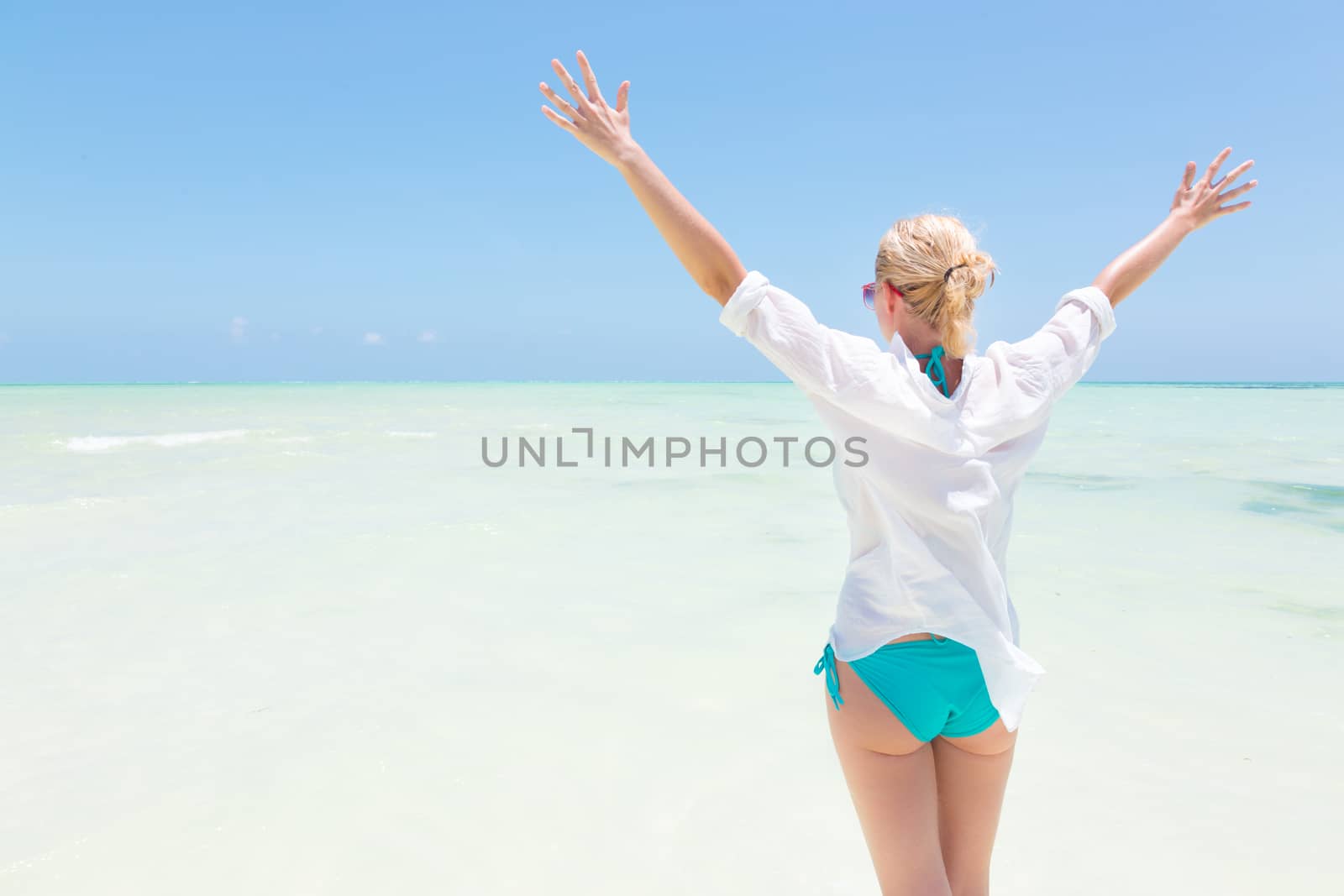 Happy woman, arms rised, enjoying, relaxing joyfully in summer at white tropical beach. Beautiful caucasian model wearing white beach tunic on vacations on Paje beach, Zanzibar, Tanzania.