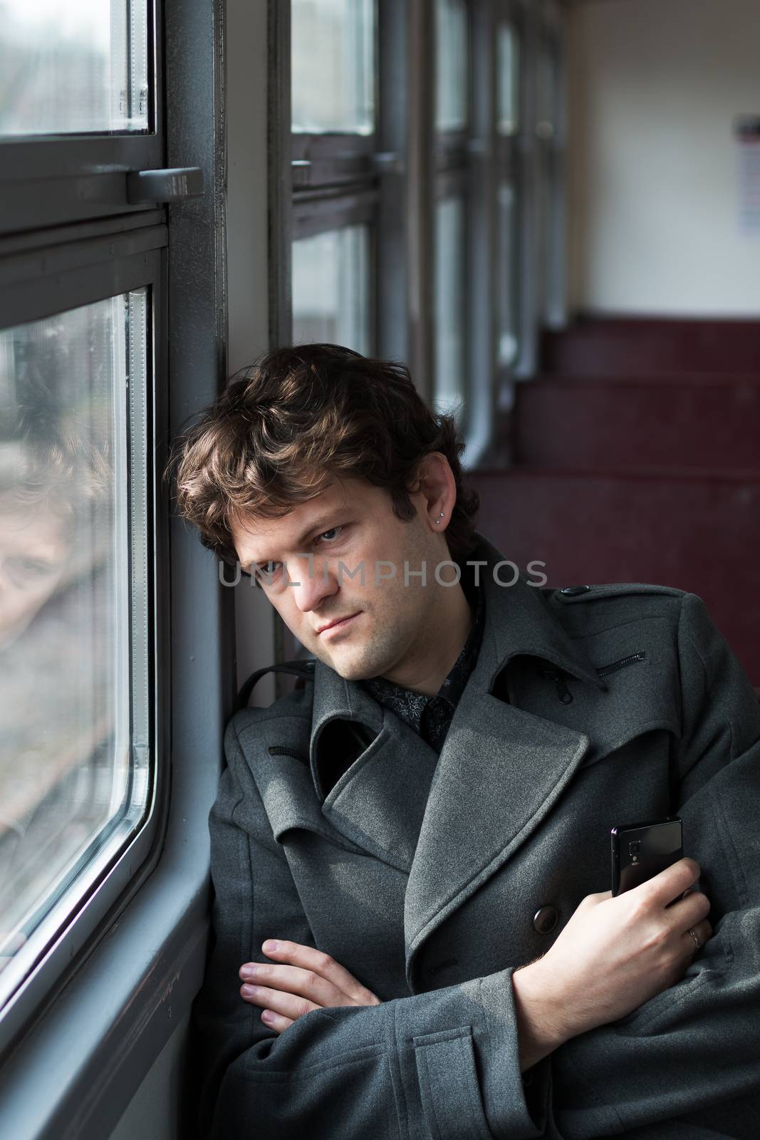 Traveling by train. Sad man traveling by train, looking through the window and thinking about unrequited love squeezing the phone in his hand. alone in an empty train wagon. Vertical. by MSharova