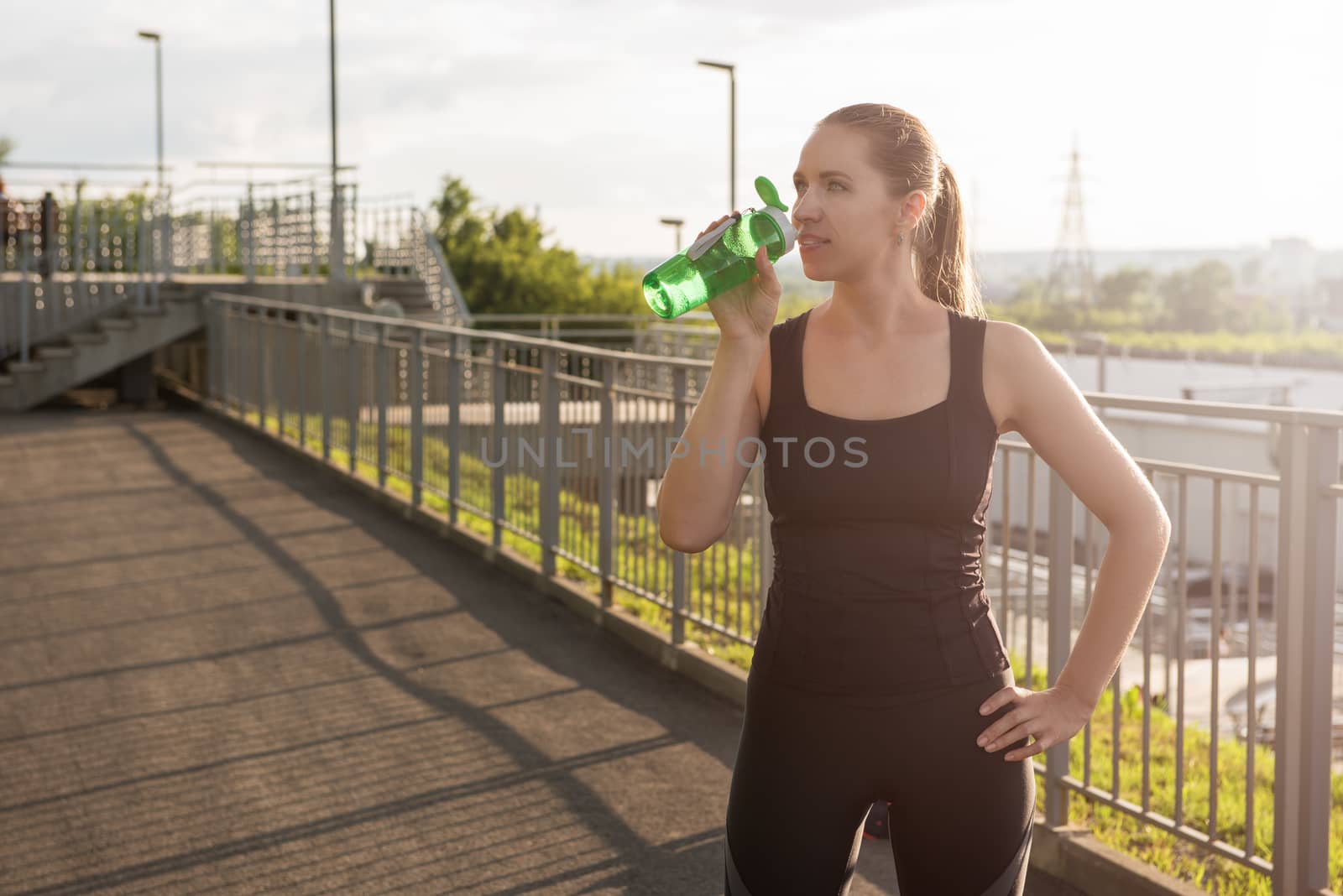 A woman in sportswear with bottle of water have rest at sunset on the evening city background