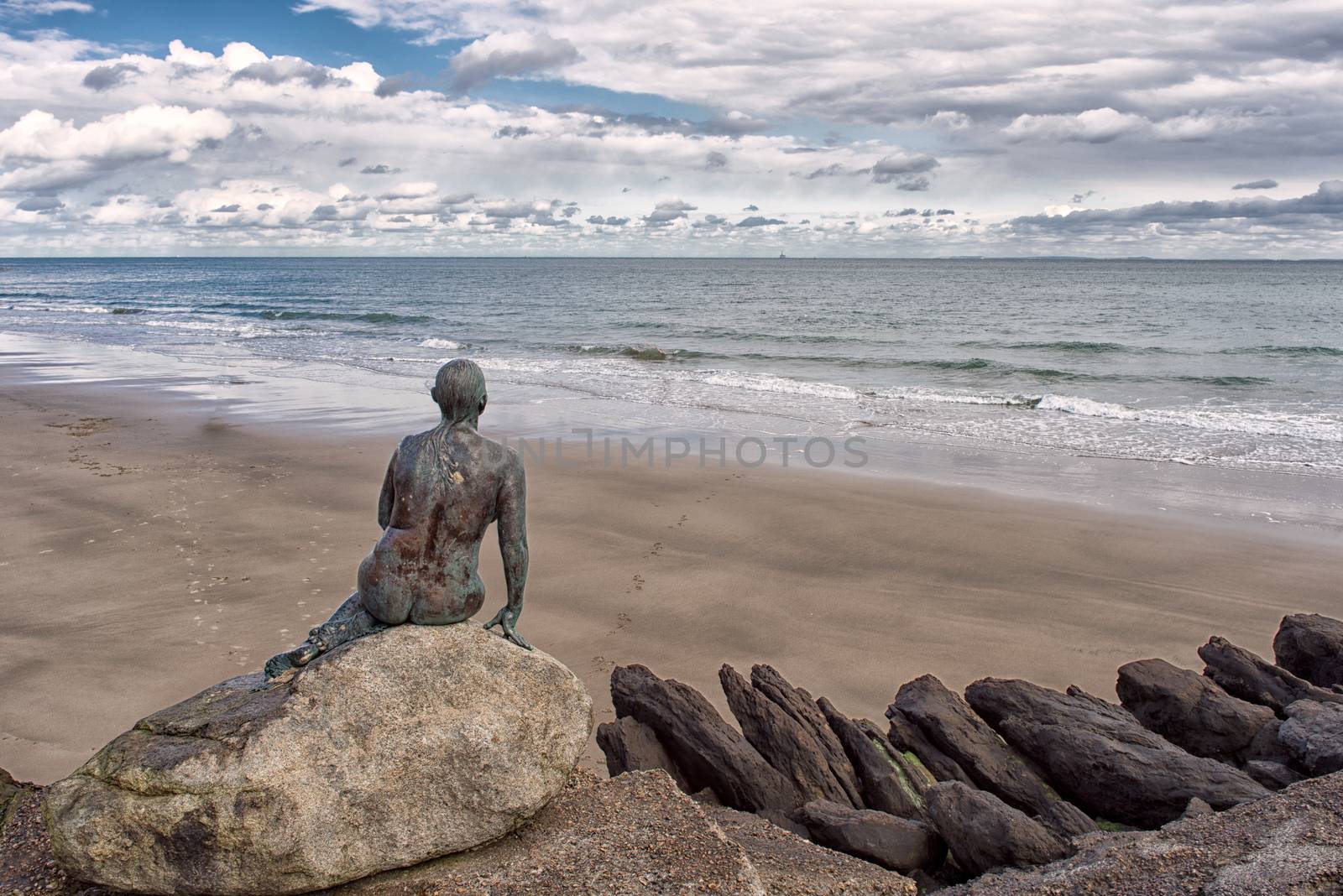 A bronze statue sitting on rocks in Folkestone overlooking a deserted beach and looking out to sea