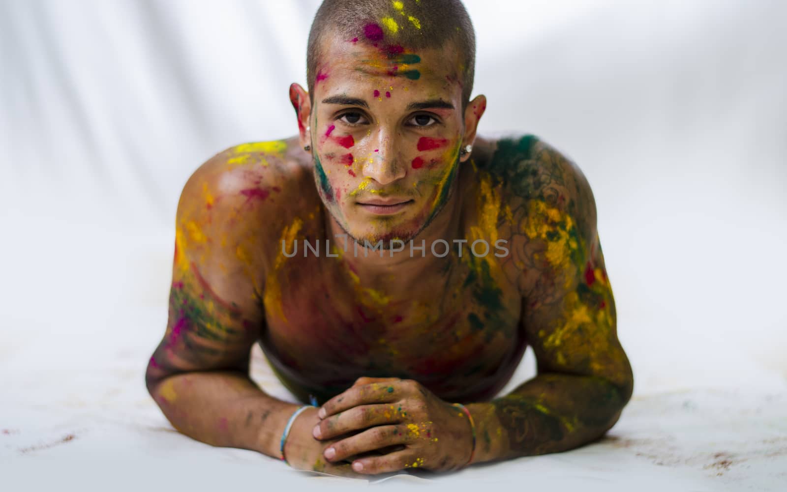 Attractive young man shirtless, skin painted all over with bright Holi colors, looking at camera, laying on the floor on white background