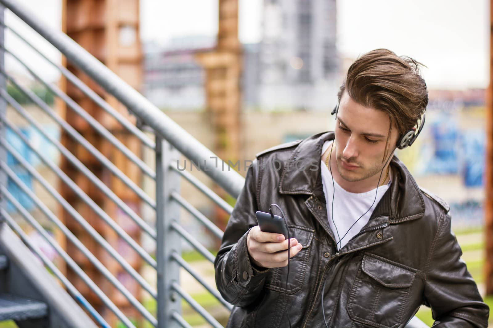 Handsome young man standing outdoors in urban environment on metal stairs, using cell phone