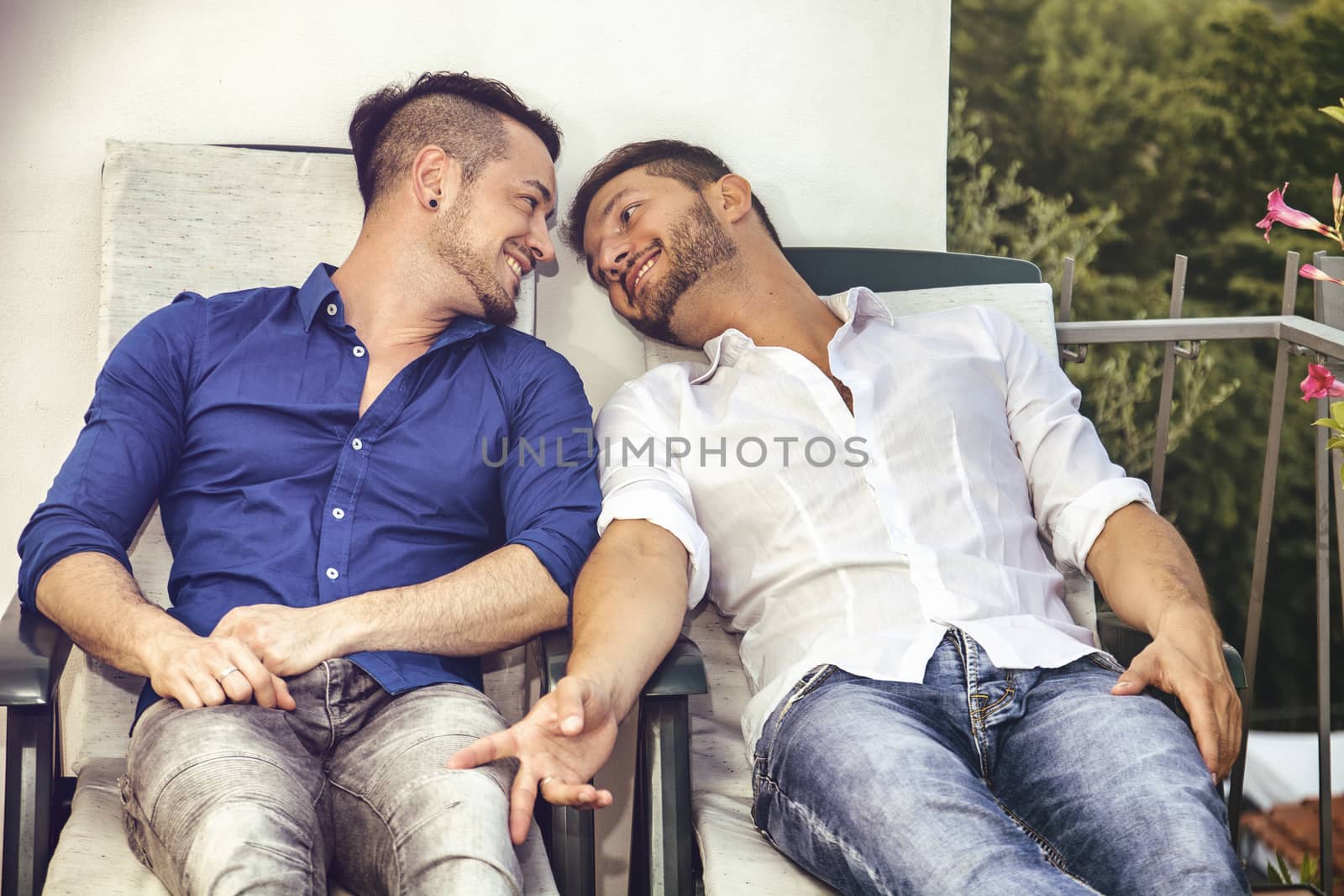 Two gays sitting on chairs at balcony holding hands and looking at each other.