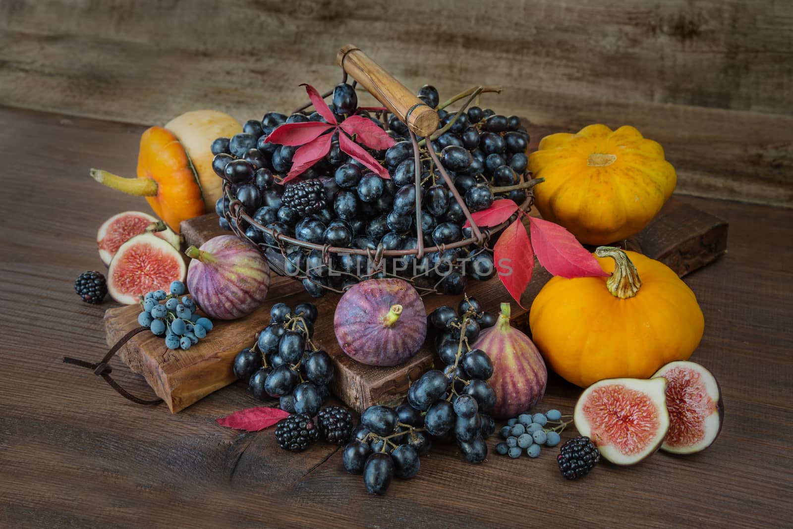 Beautiful autumn still life: black grapes in a basket on the old cutting board as well as ripe figs, red raspberries, orange pumpkin and blackberries with autumn leaves on the wooden background