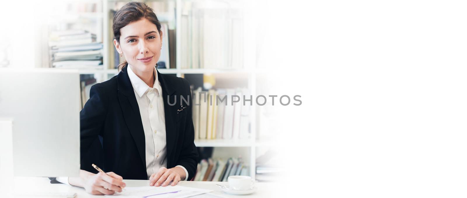 Portrait of smiling businesswoman looking at camera and smiling while working with documents at office in front of computer