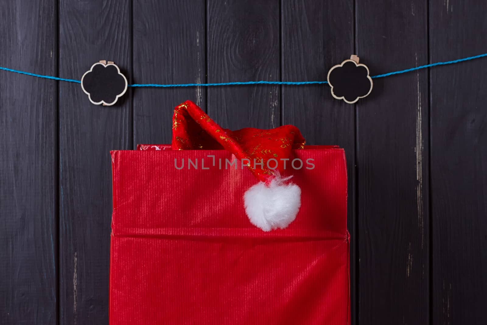 large red box from which the cap of Santa Claus sticks out against the background of a wooden black fence. Christmas