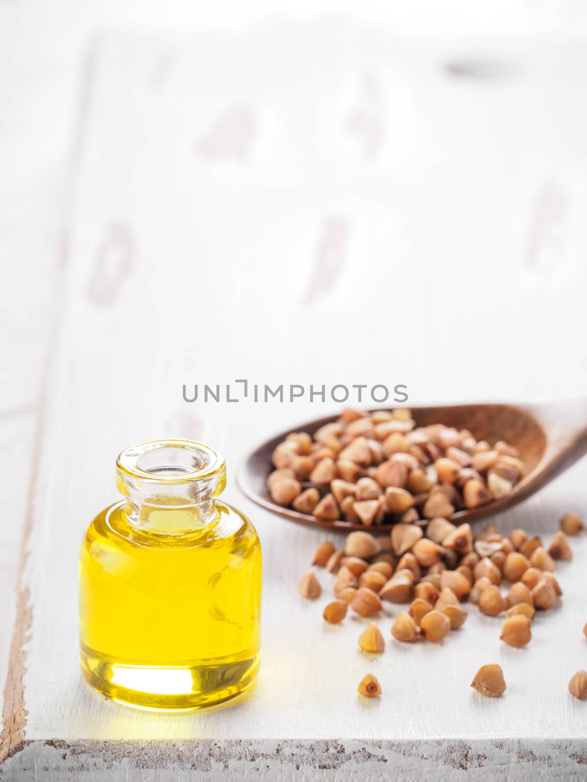 Brown buckwheat in spoon and buckwheat oil in glass bottle on white wooden background. Copy space.