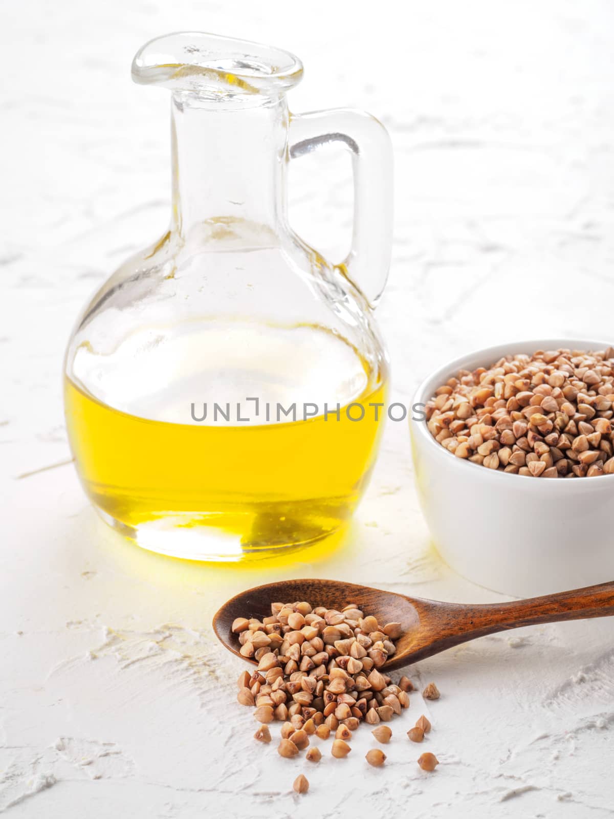Brown buckwheat in spoon and buckwheat oil in glass bottle on white wooden background. Copy space.