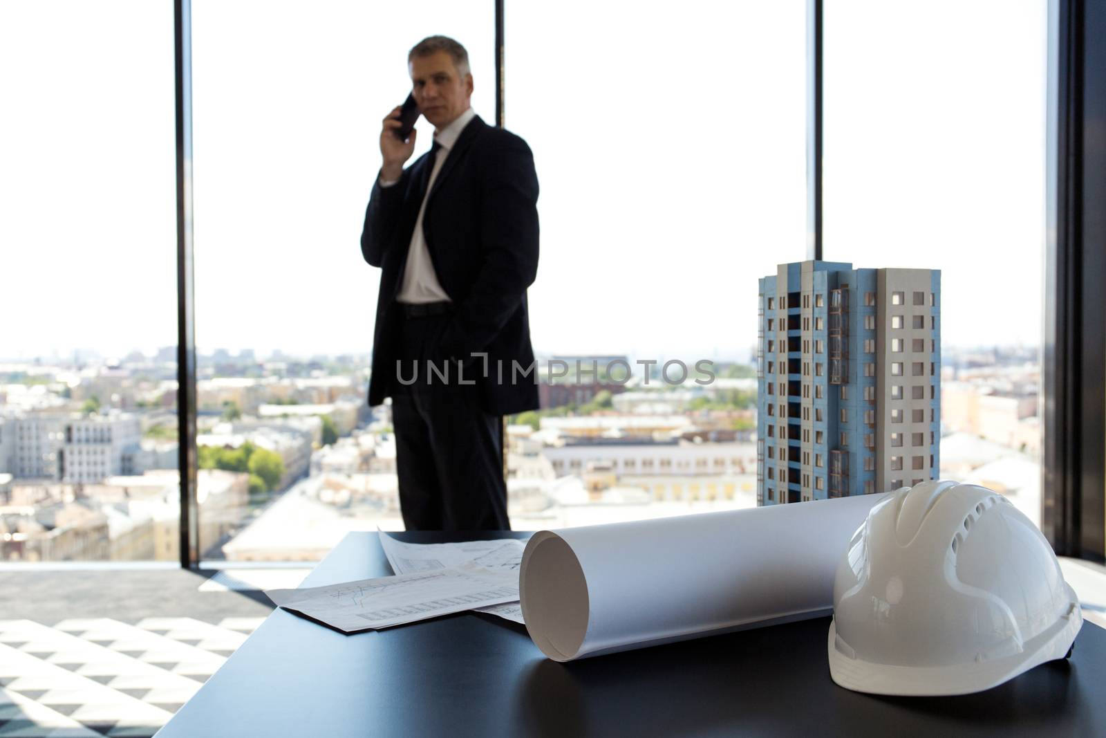 Businessman in construction company office, model of modern multi storey residential building house, hardhat, blueprint and documents on table