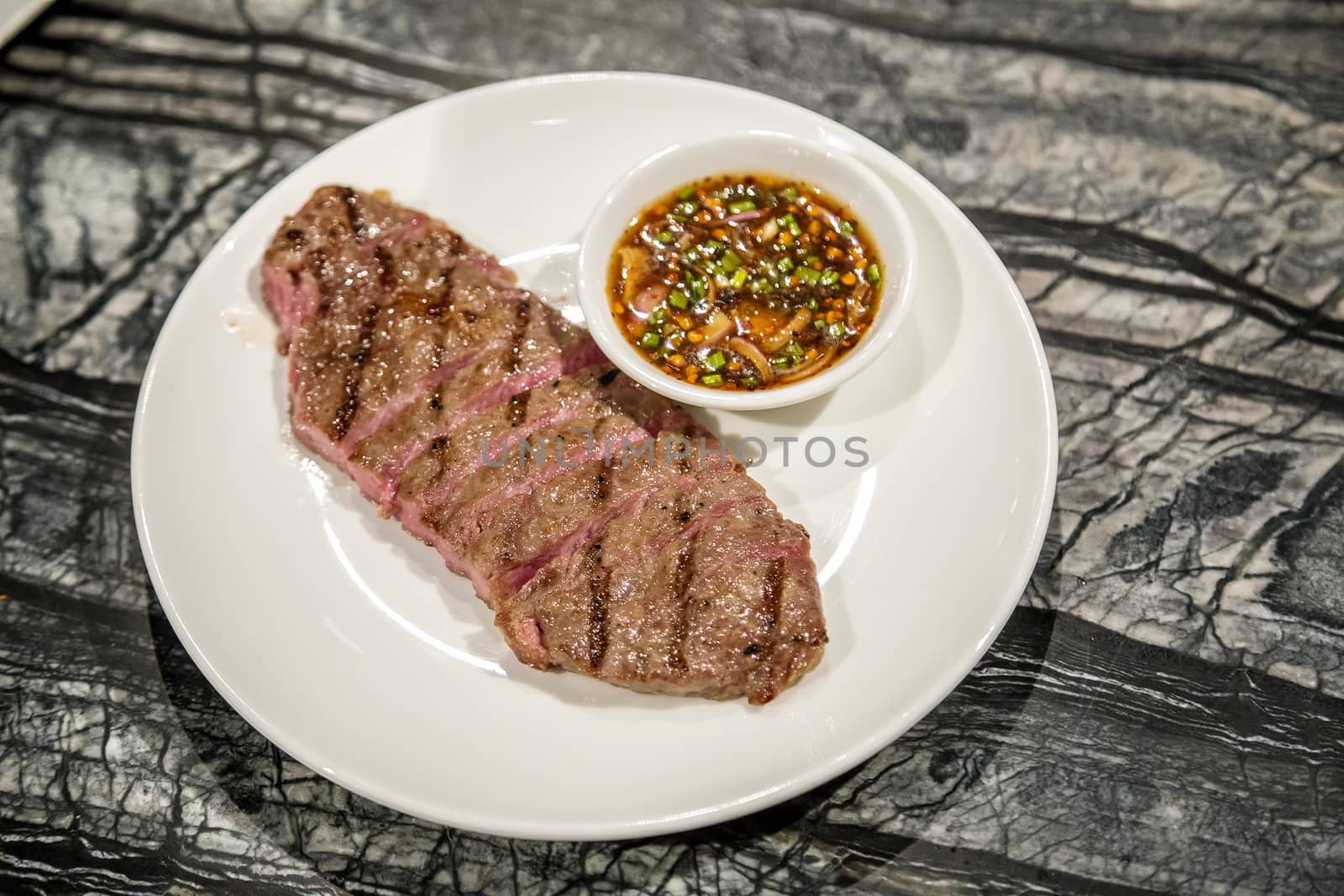 Grilled beef steak with Thai style spicy sauce on white plate and granite table, Medium rare barbecue steak, Thai style food.