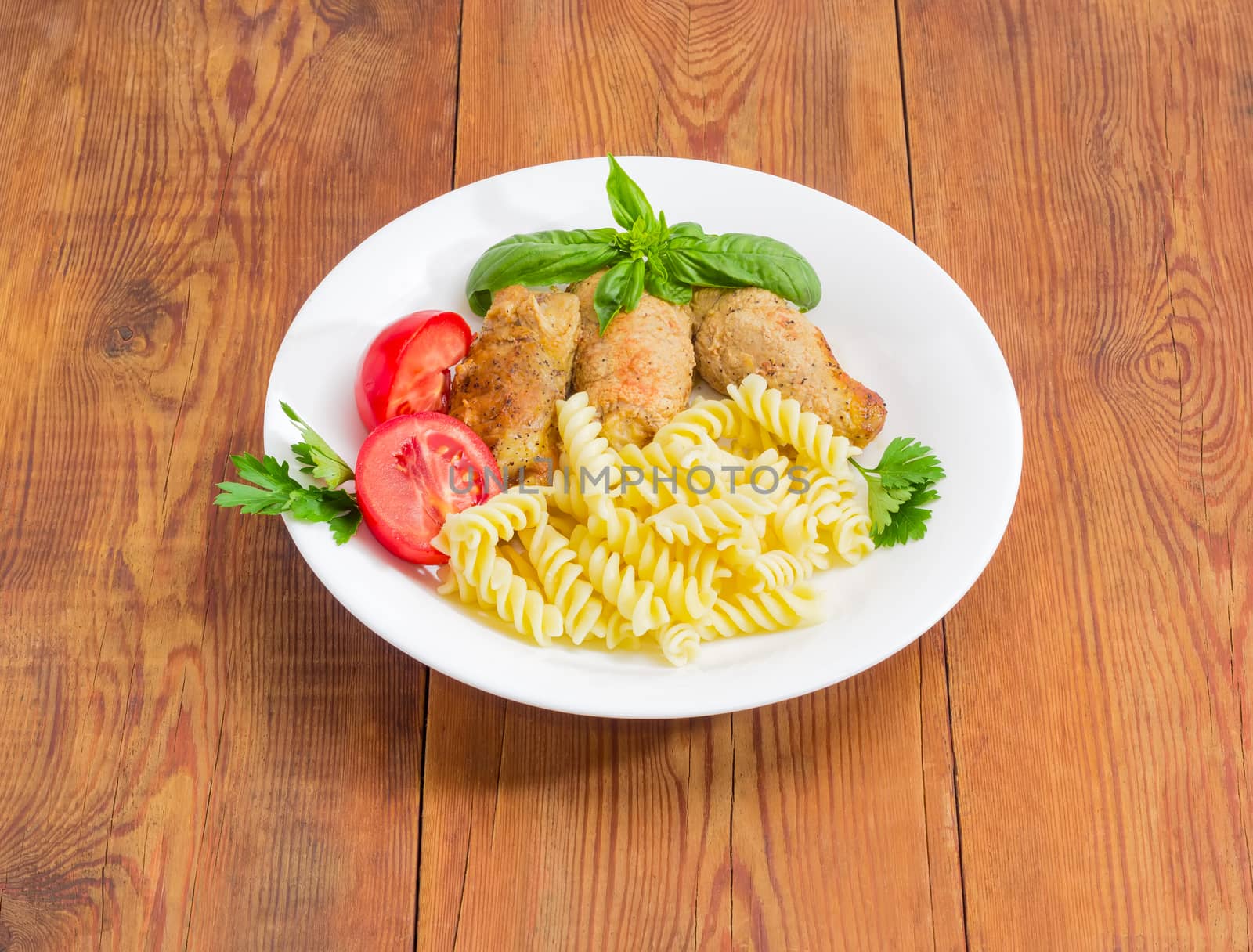 Braised meat roulades with filling from bread dried crusts and mushrooms, tomato and spiral pasta decorated with basil and parsley twigs on a white dish on a surface of an old wooden planks
