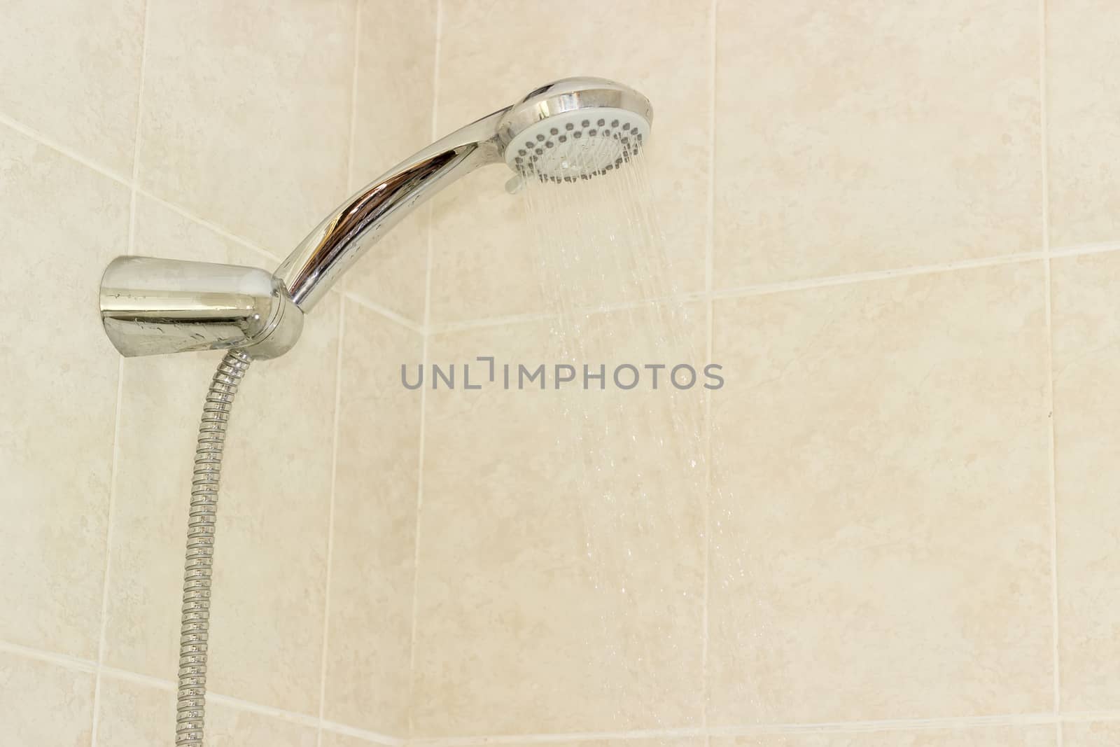 Water flowing from shower head with turn lever of a spray settings and metal shower hose in holder on a background of a wall with beige tiles
