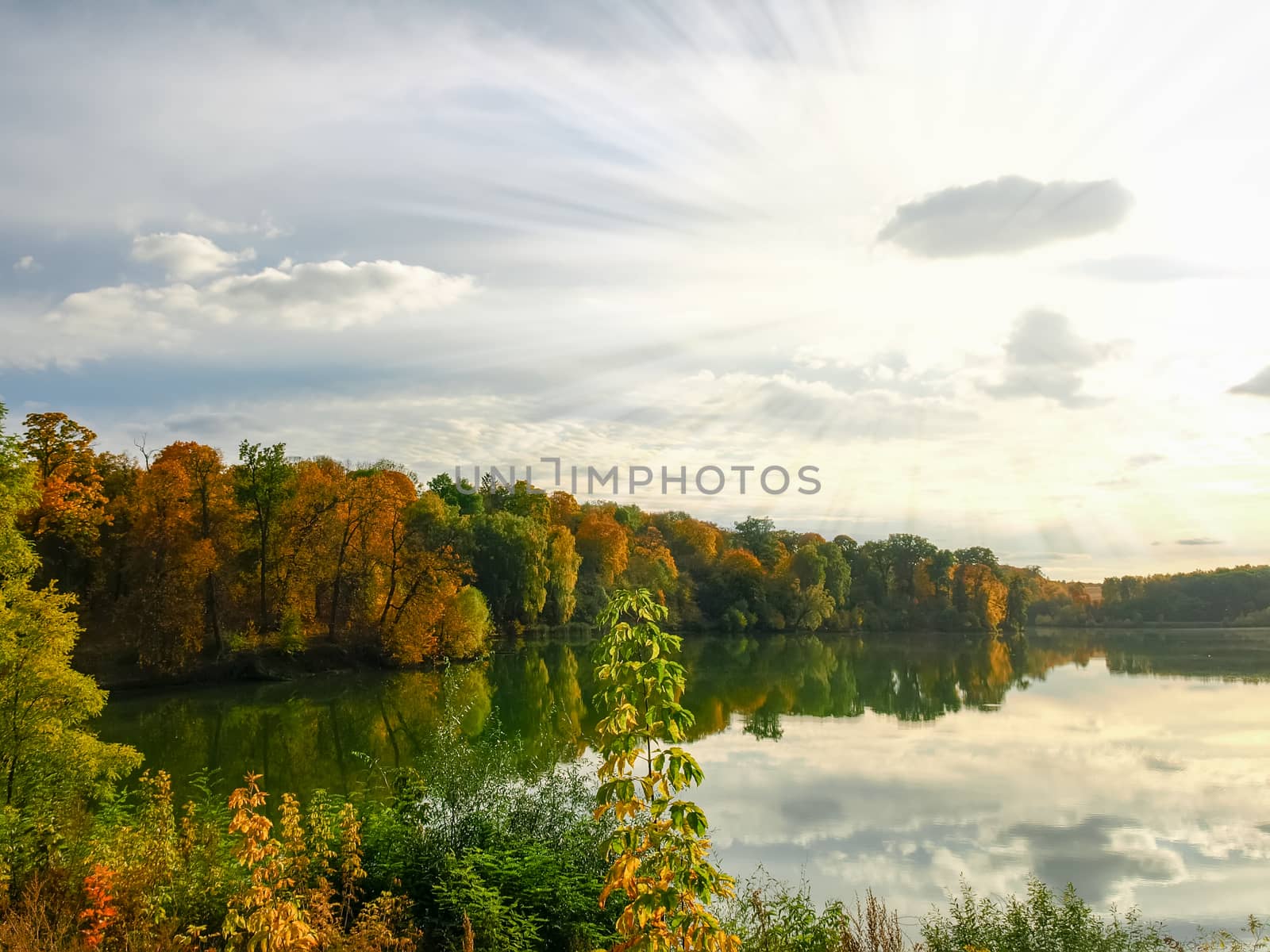 Autumn forest on the shore of the lake and reflection of trees and sky with clouds in calm surface of the water at sunset
