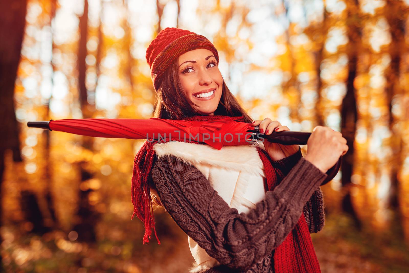 Portrait of a beautiful smiling girl with red umbrella in sunny forest in autumn colors. Pensive looking away.