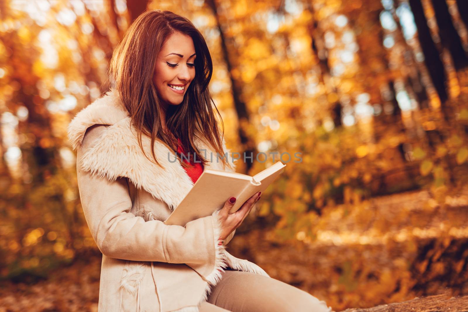 Beautiful young smiling woman reading book in sunny forest in autumn colors.
