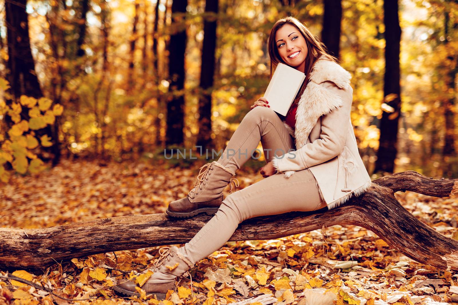 Beautiful smiling girl enjoying in forest in autumn. She is sitting on the tree, holding book and thinking.