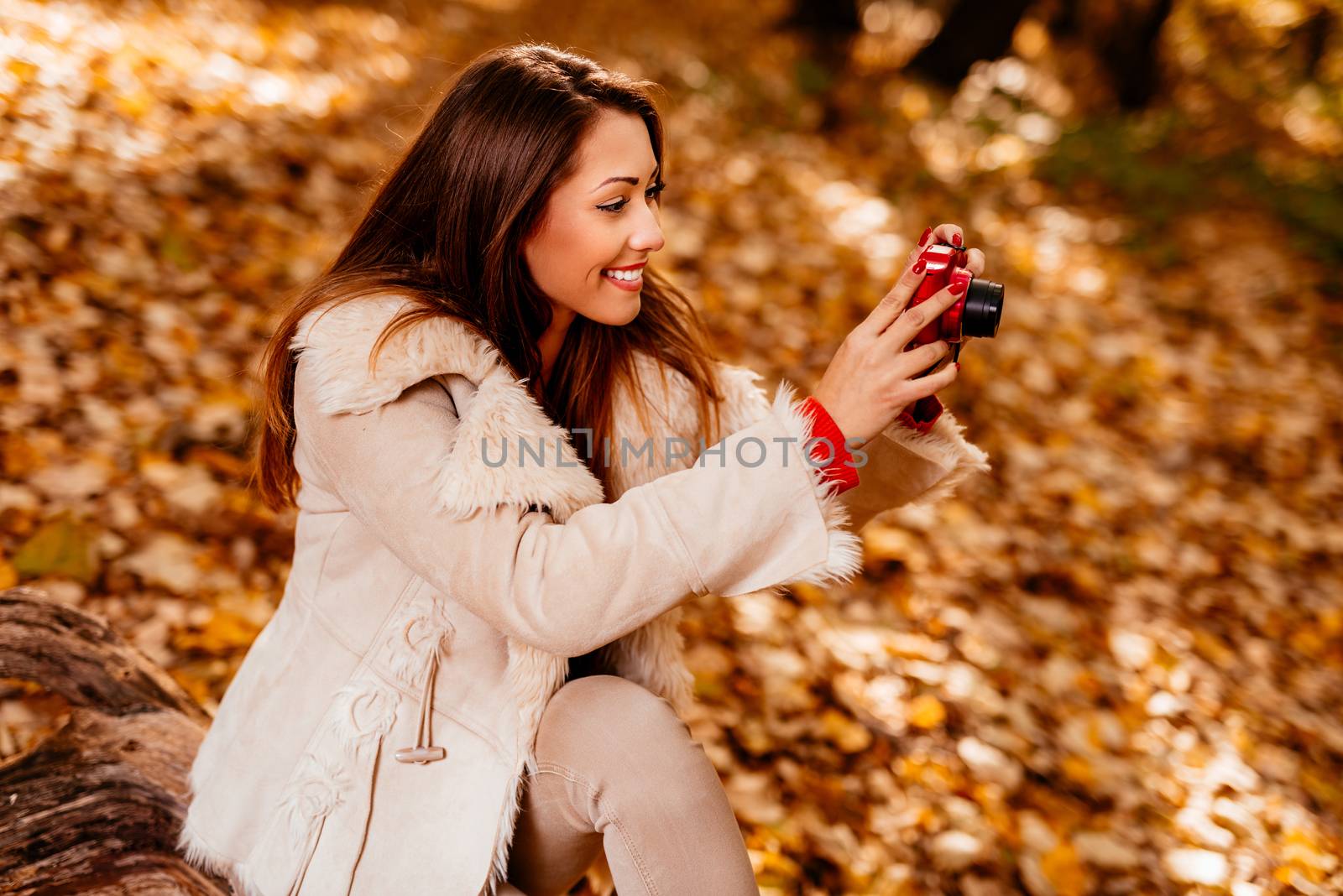 Beautiful smiling girl enjoying in forest in autumn. She is sitting on the tree and taking photo for memory.