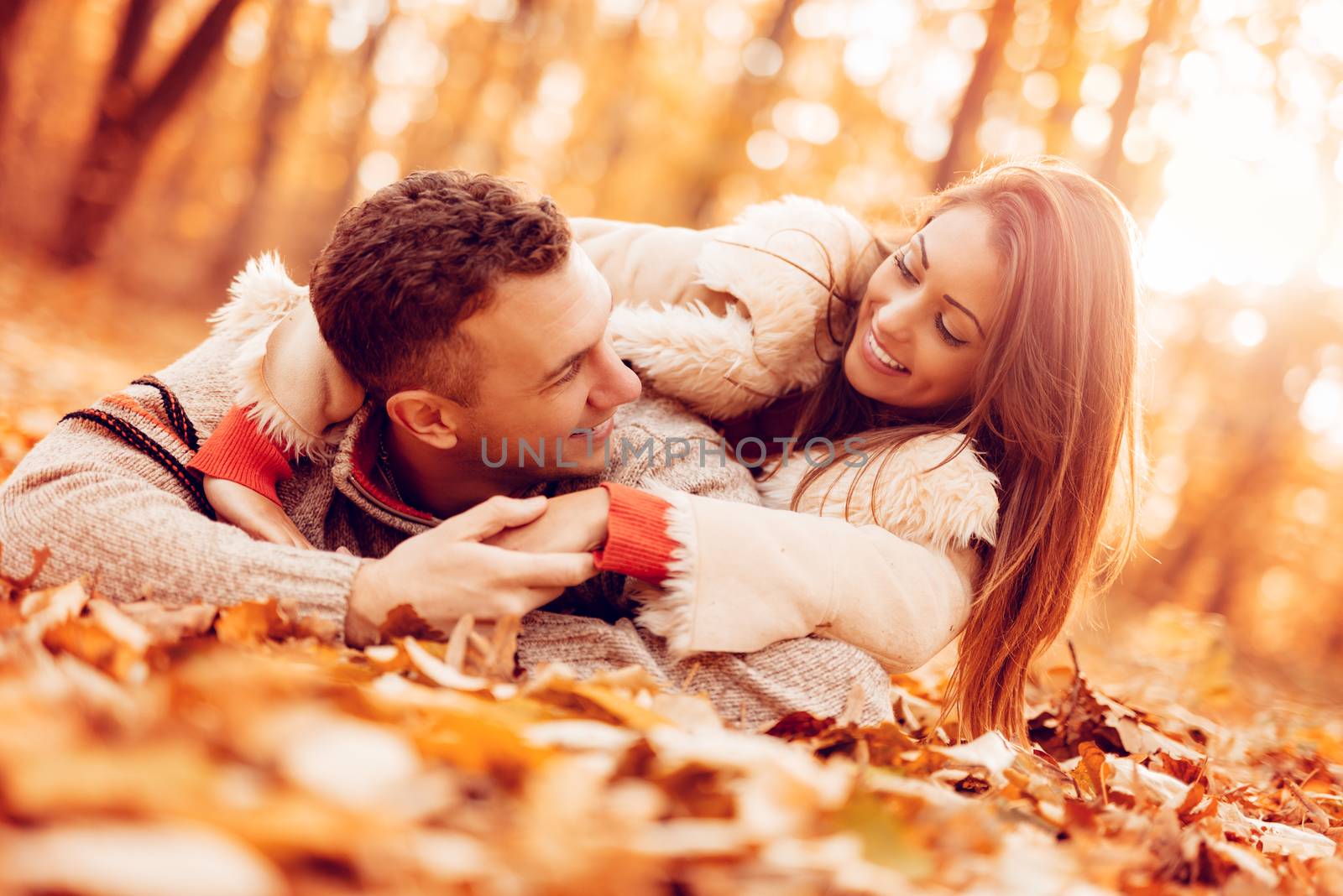 Beautiful smiling couple enjoying in sunny forest in autumn colors. They are lying on the falls leaves and having fun.