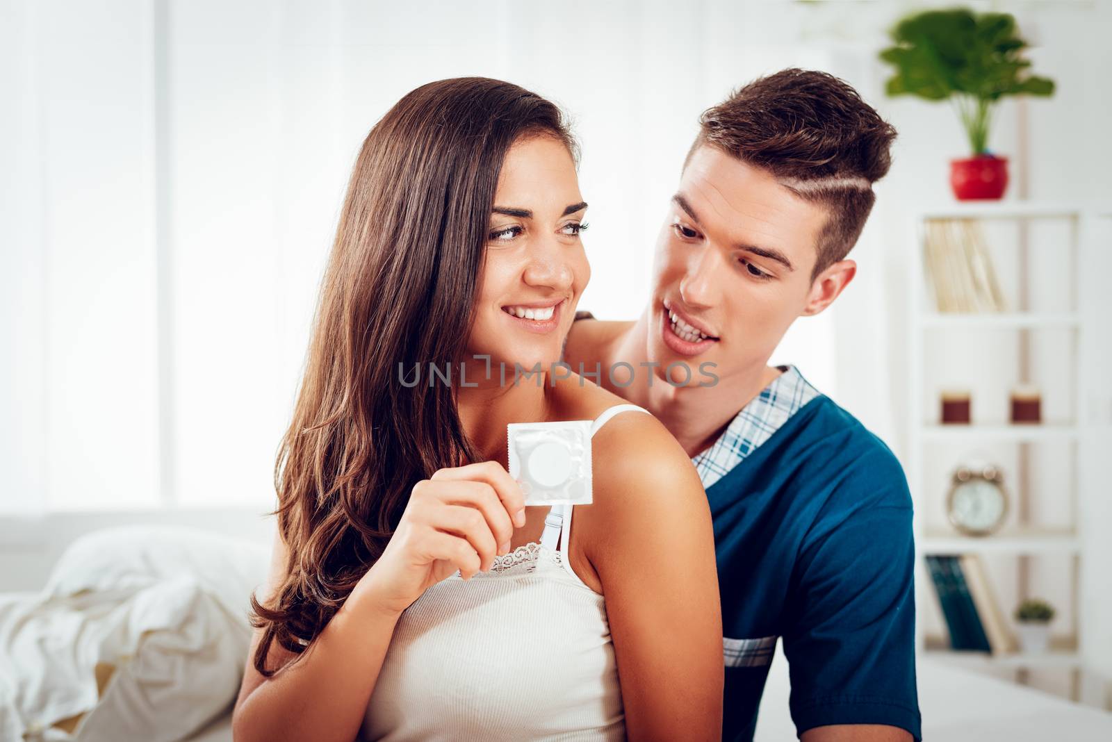 Young couple sitting in bed looking at each other while a girl holding a condom.
