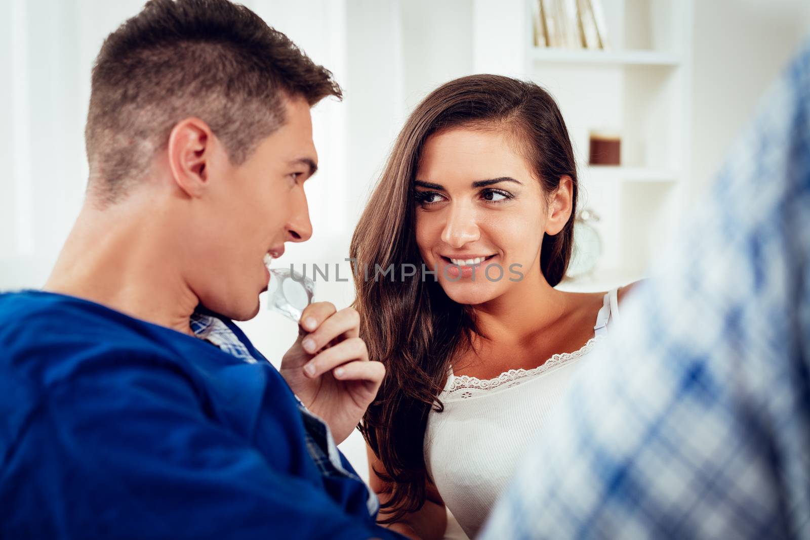 Young couple lying in bed looking at each other while a guy holding a condom in his mouth.