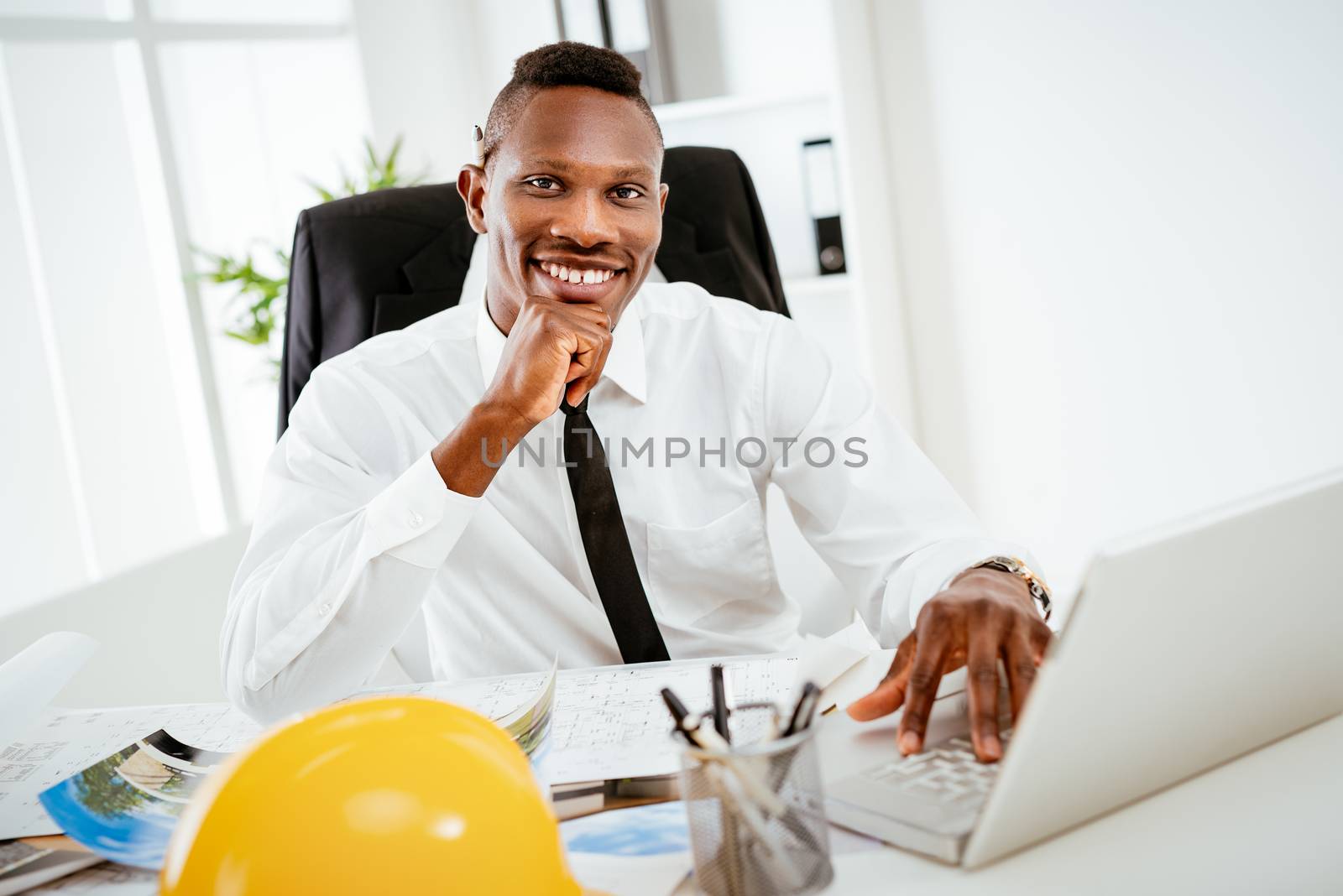 Smiling African construction engineer sitting at desk in the office and looking at camera. 