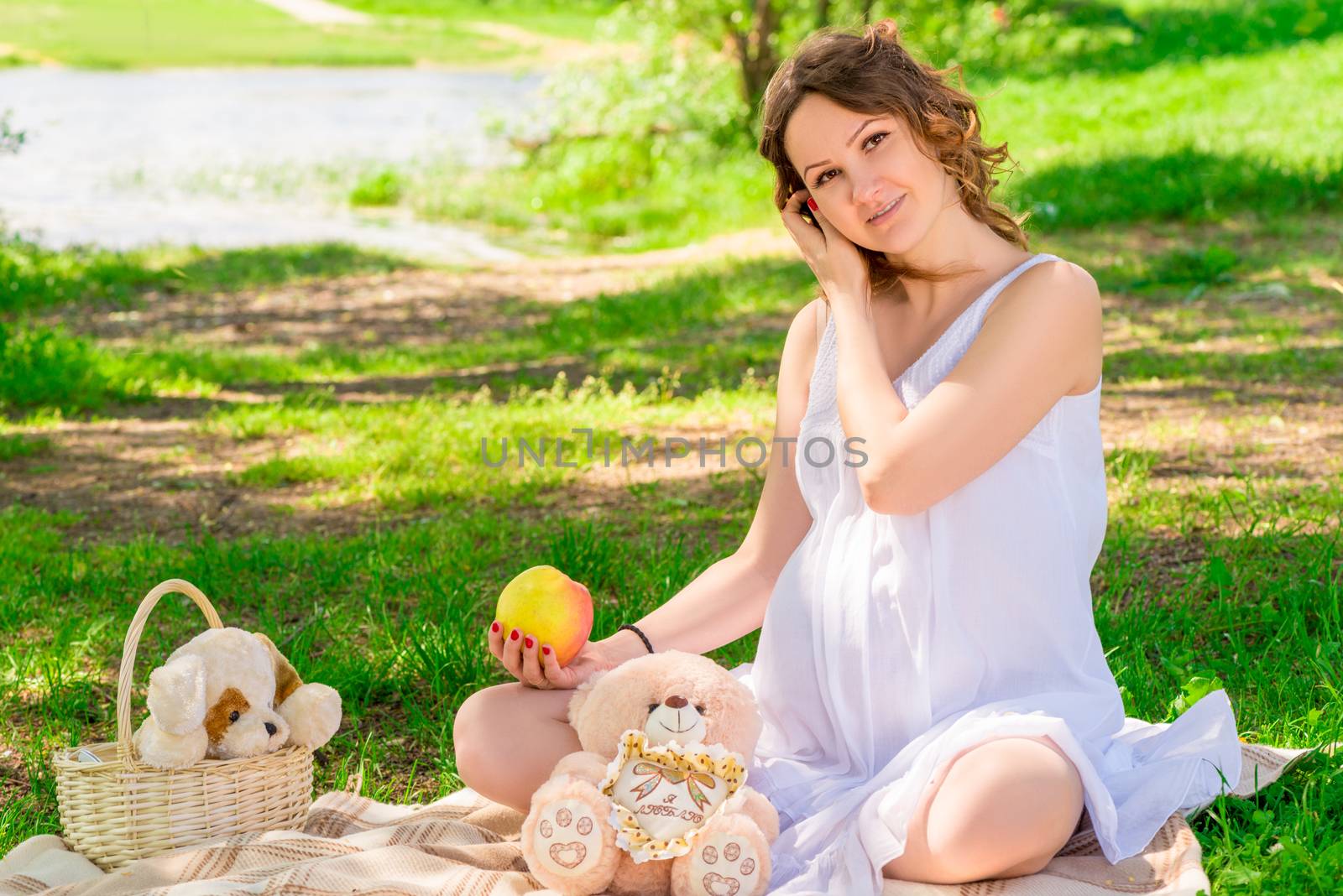 Young pregnant girl in a white sarafan on a plaid in a park at a picnic