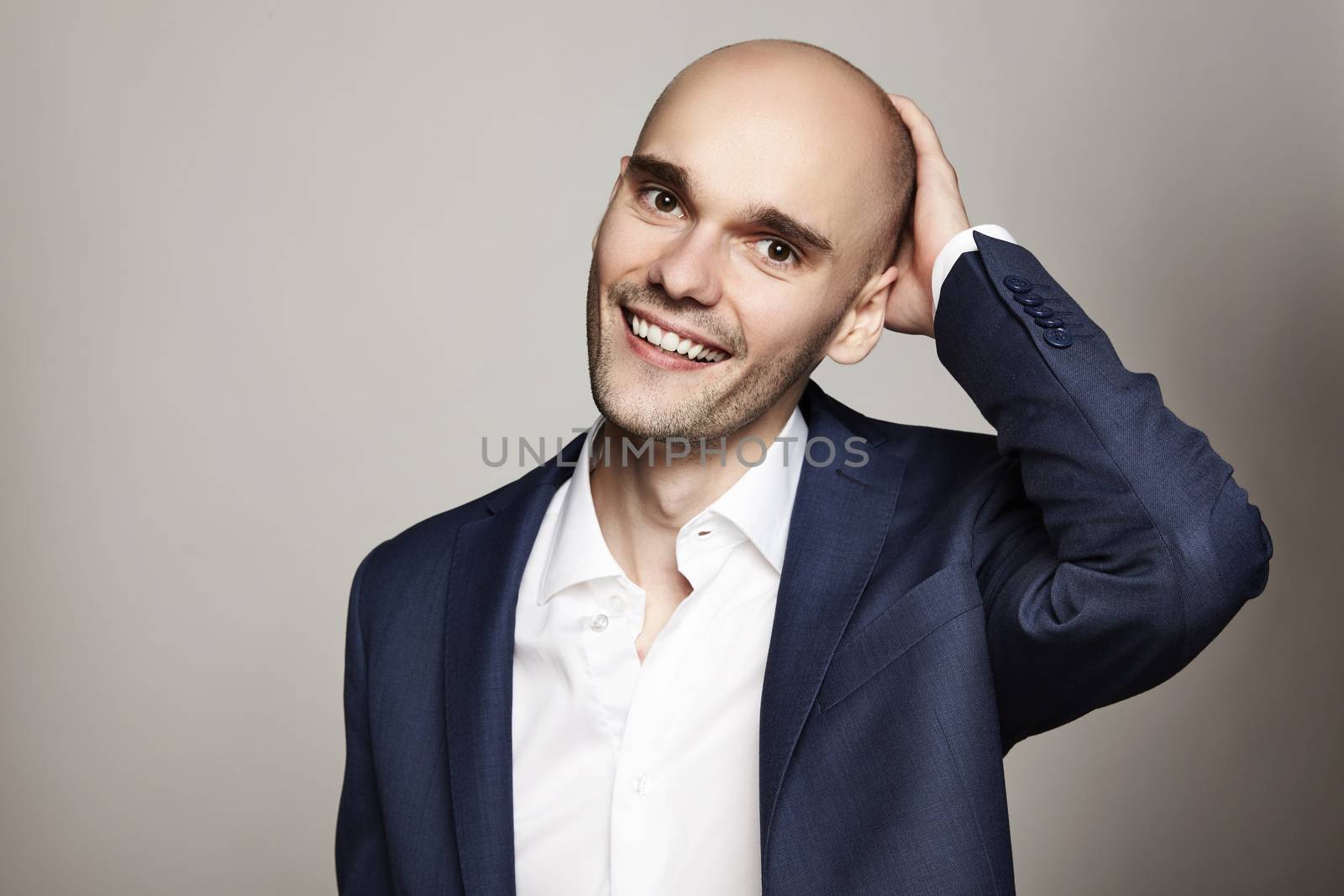 Close-up portrait of a young bald man stroking his head. He is smiling. Gray background. Horizontal.